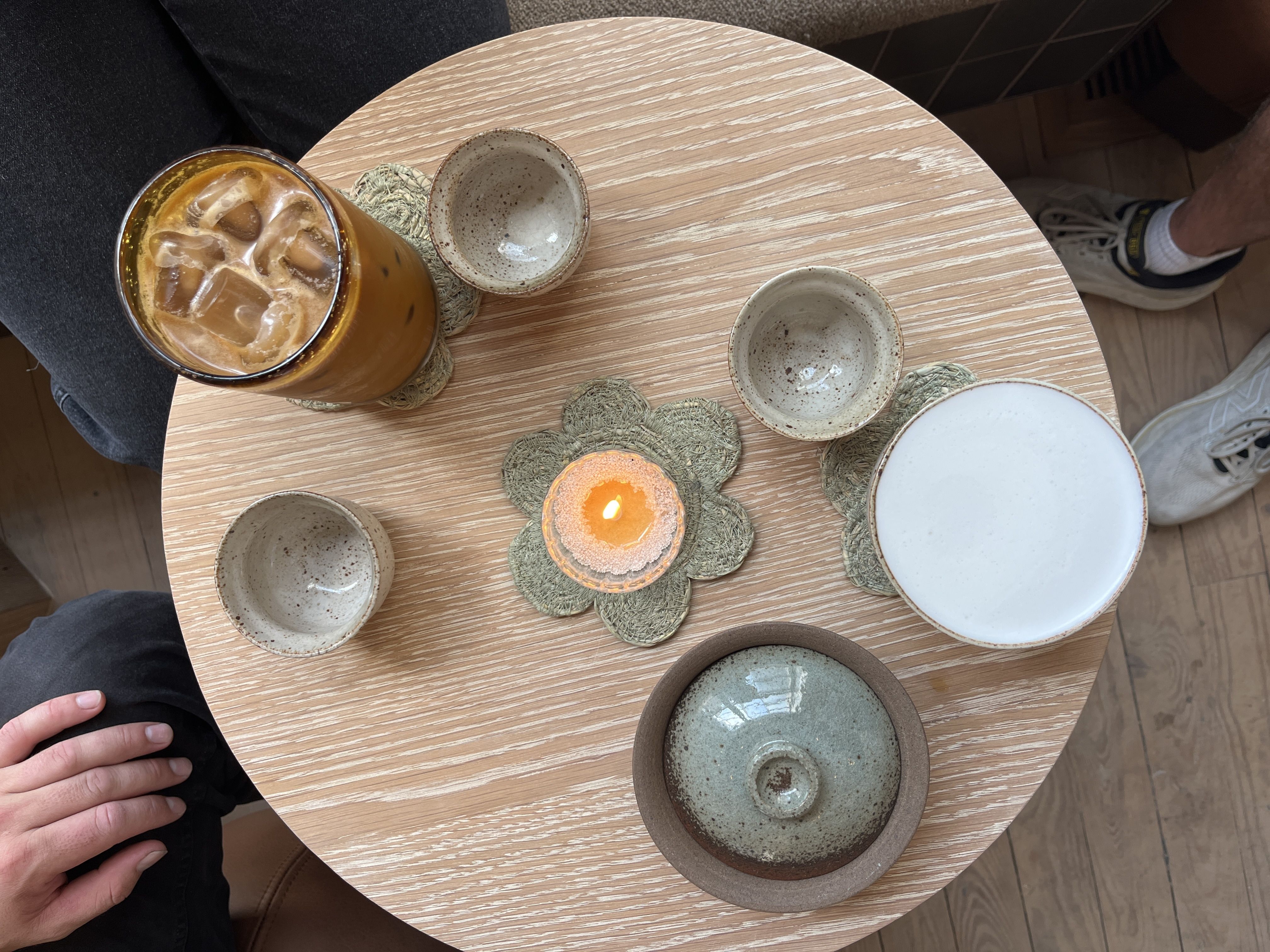 Round wooden table with iced coffee, ceramic cups, a lidded bowl, a drink with foam, and a lit candle on flower-shaped coasters, with two people seated nearby.
