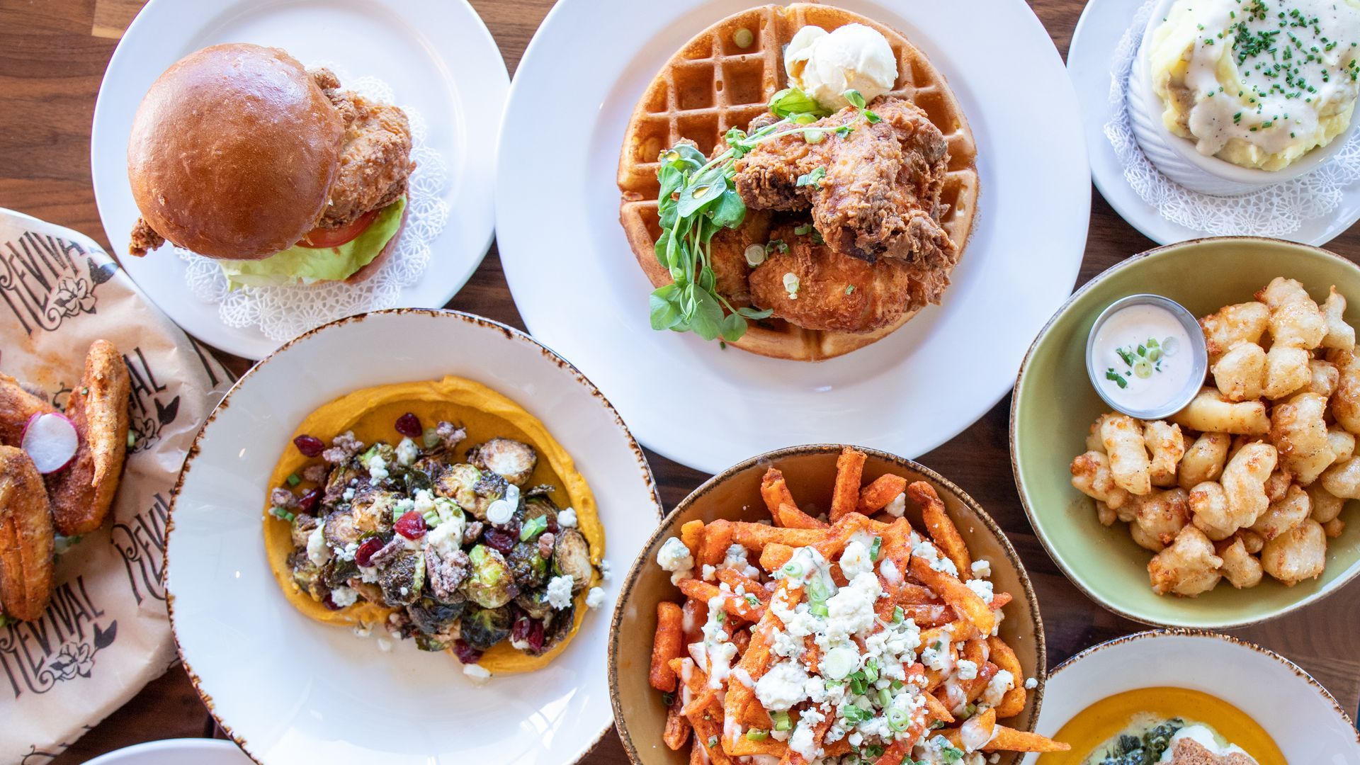 Top-down view of a colorful brunch spread: crispy fried chicken on a waffle, a burger on a bun with lettuce, bowls of loaded toppings and potatoes, plus mashed potatoes with chives.
