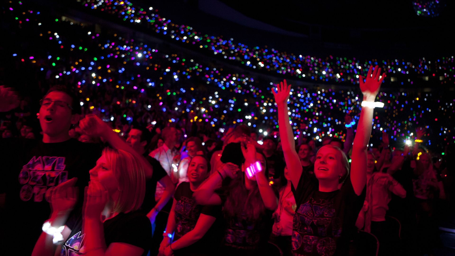 Nighttime concert scene: a packed crowd with thousands of colorful LED wristbands and phone lights, hands raised in excitement as red stage lighting washes the foreground.