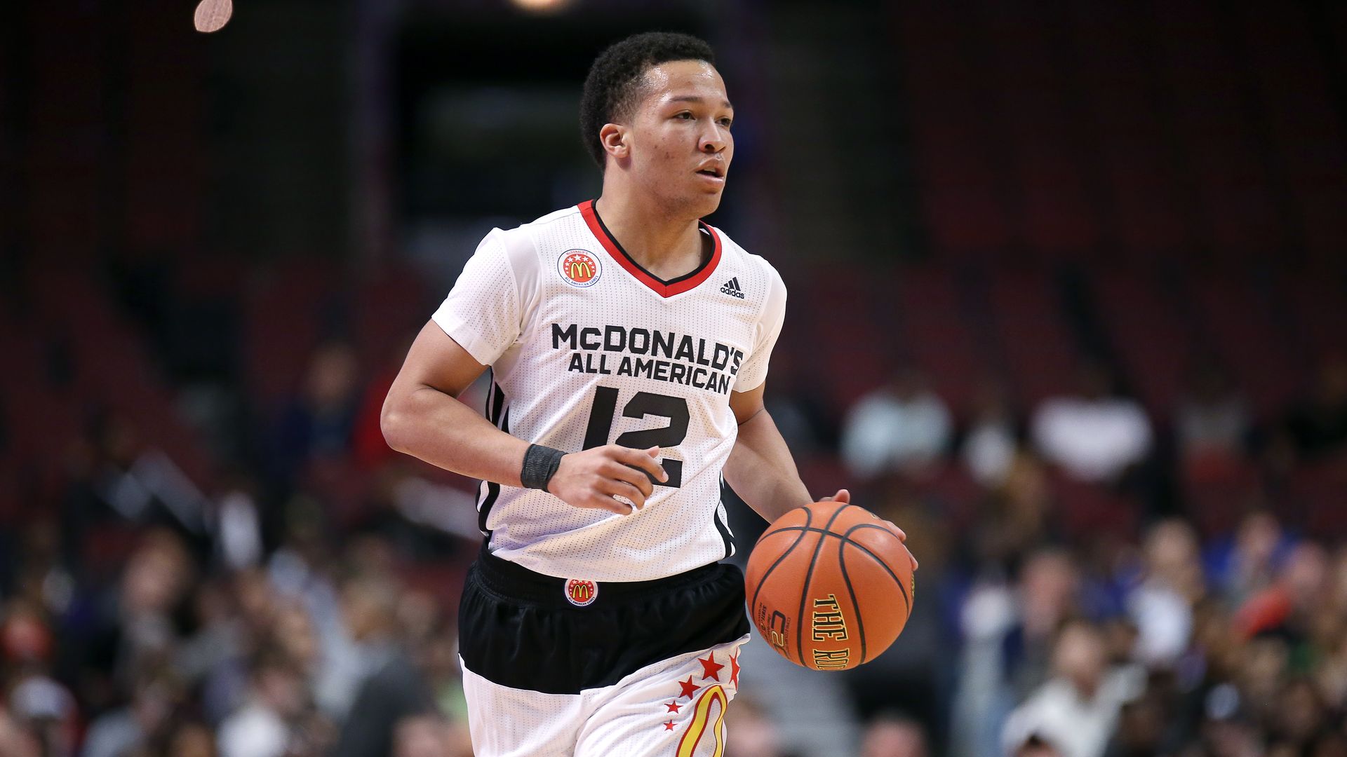 A basketball player in a white McDonald's All American jersey (number 12) dribbles an orange ball on a court, with a blurred crowd in the stands behind him.