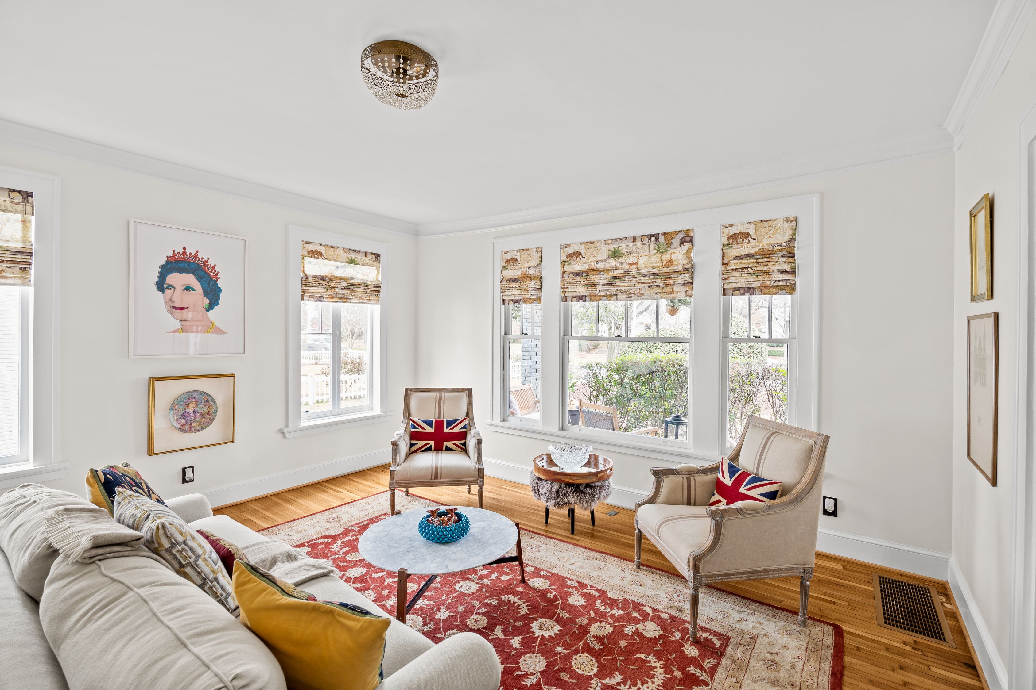 Bright living room with beige sofa, two beige armchairs with Union Jack pillows, red floral rug, round coffee table, animal print window shades, and portrait of a woman wearing a red crown.