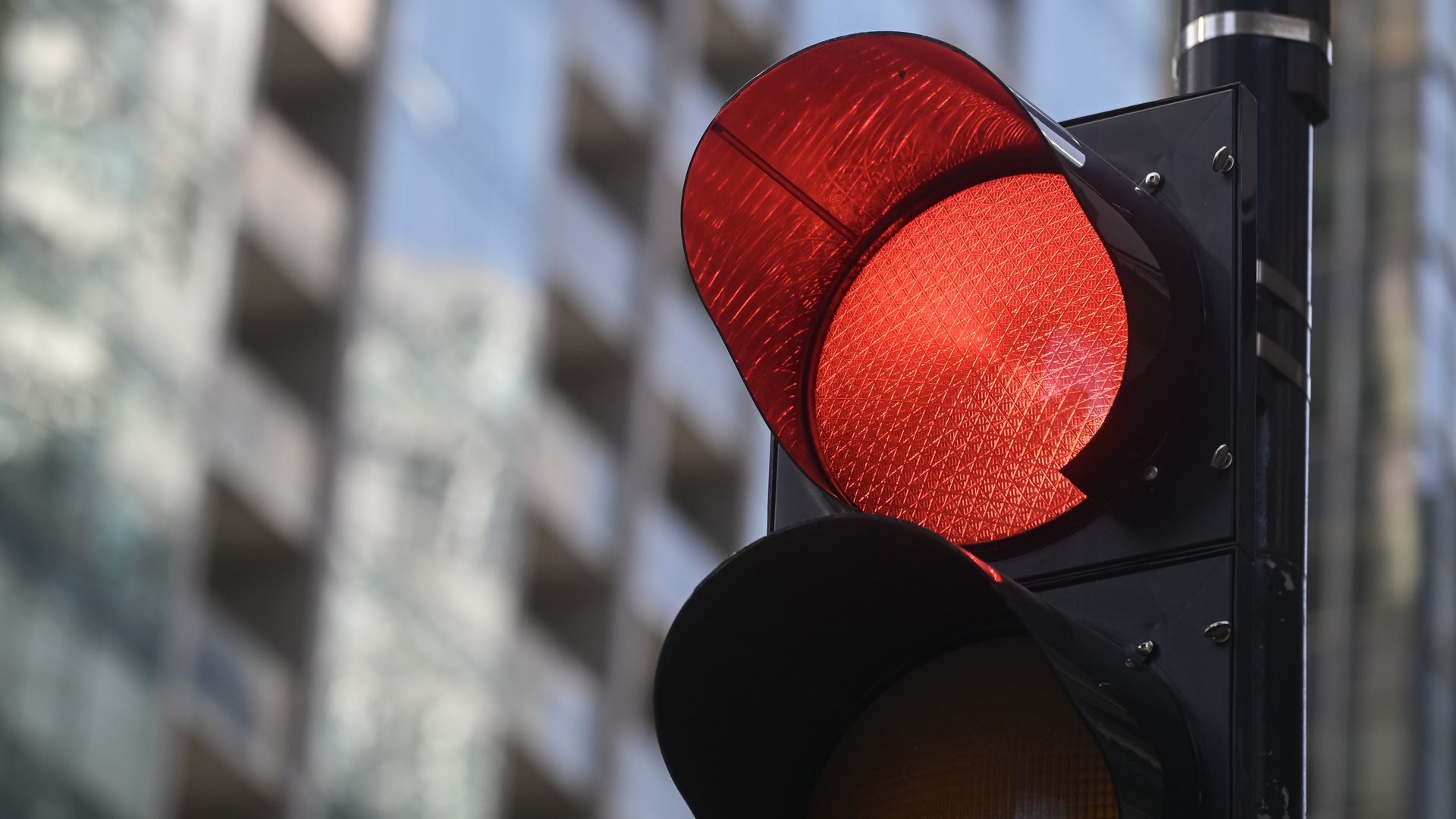 Close-up of a red traffic light on a black pole, featuring a circular textured lens with a geometric pattern; a blurred urban street background.
