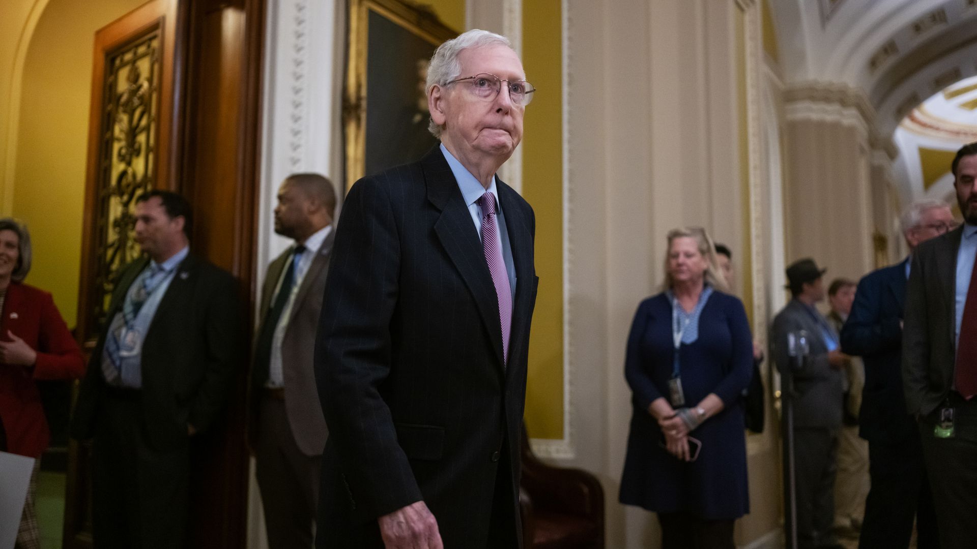 Senate Minority Leader Mitch McConnell, wearing a dark gray suit, light blue shirt and purple tie, walking out of the Senate chamber.