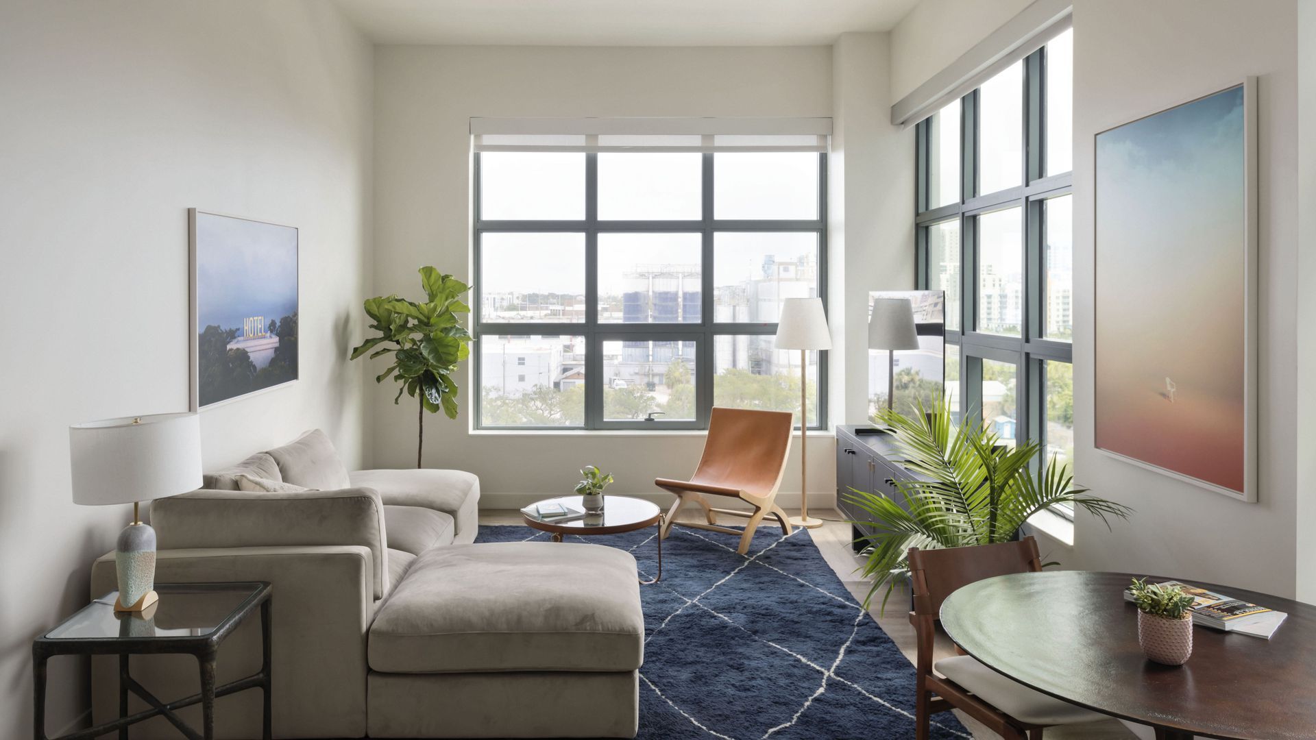 A living room with modern furniture, a blue rug, a painting on the wall and large windows.