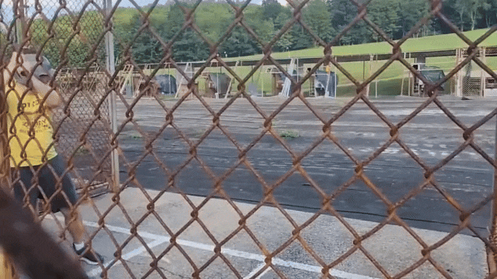View through a rusty chain-link fence of a man in a yellow shirt and black shorts hitting a baseball at a batting cage with grassy hills and trees in the background.