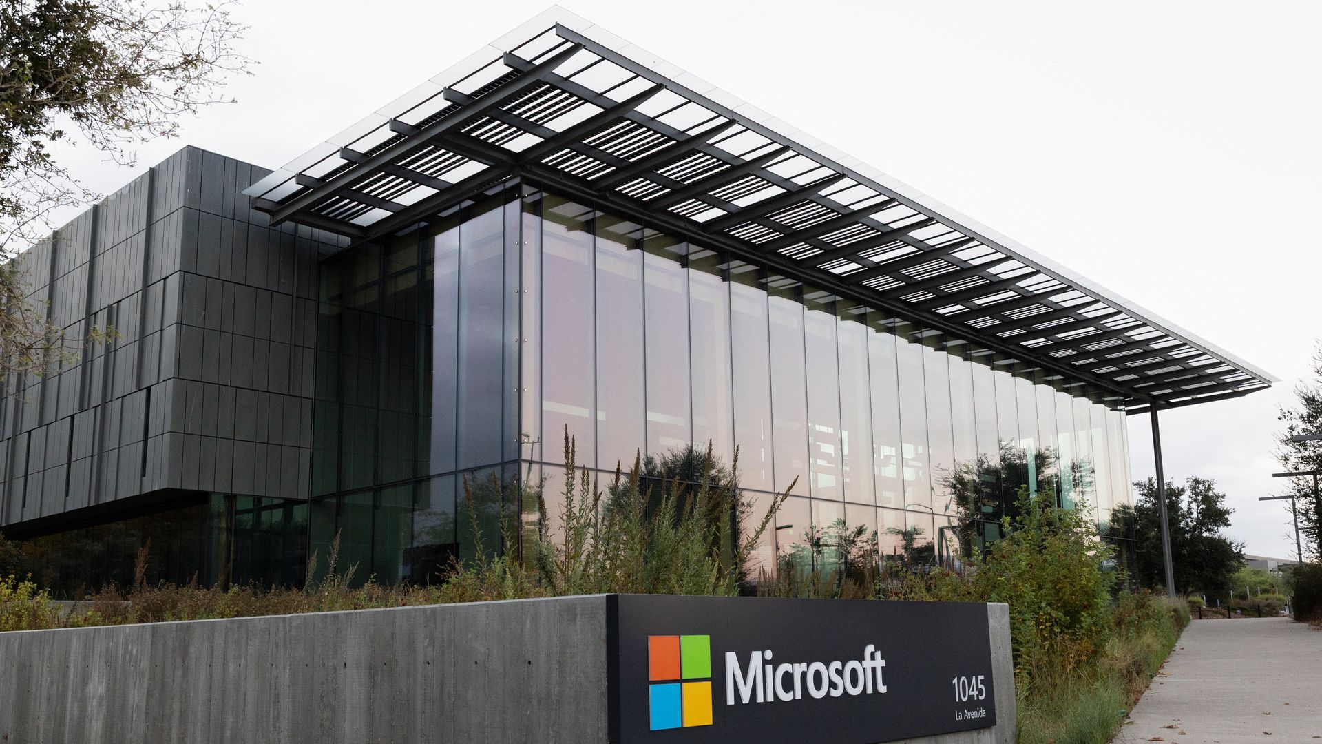 Modern glass office building with a dark steel lattice canopy over large glass windows. A concrete wall sign in front displays the Microsoft logo with colored squares and text, greenery around.