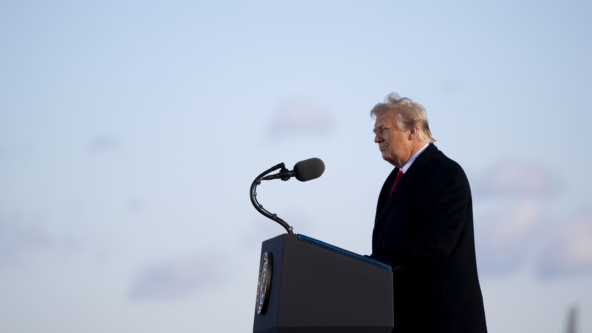 Photo of Donald Trump speaking from behind a podium, with a light blue sky in the background