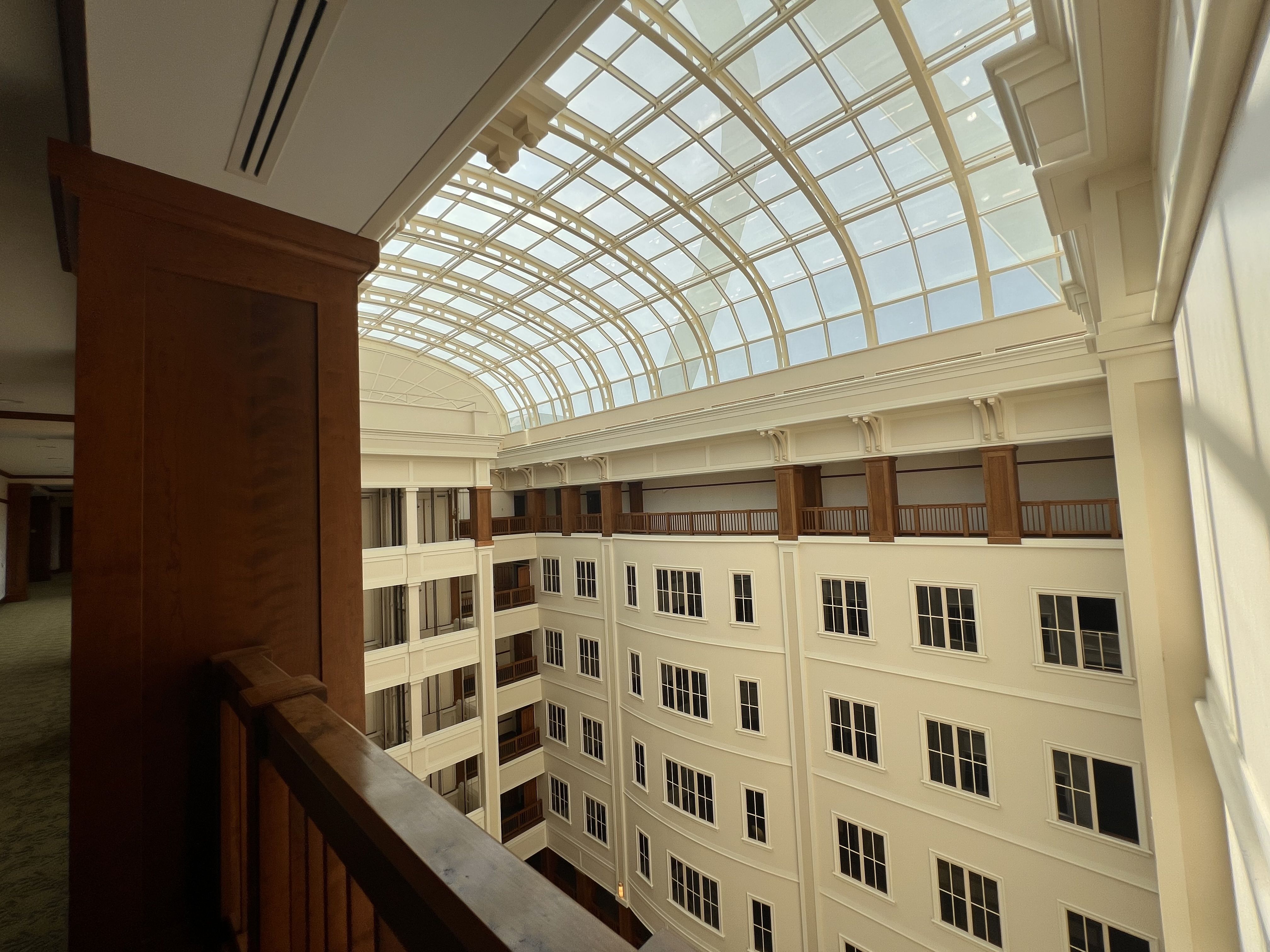 Interior atrium with a curved glass skylight and white metal arches, beige walls, and multiple levels of balconies with wooden railings surrounding a central open space.