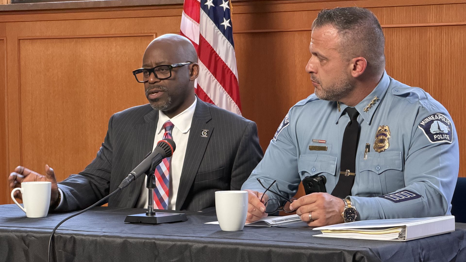 Two men sit at a panel. On the left, a bald man in a pinstripe suit and glasses speaks into a microphone; on the right, a police officer in a light blue uniform. An American flag stands behind them.