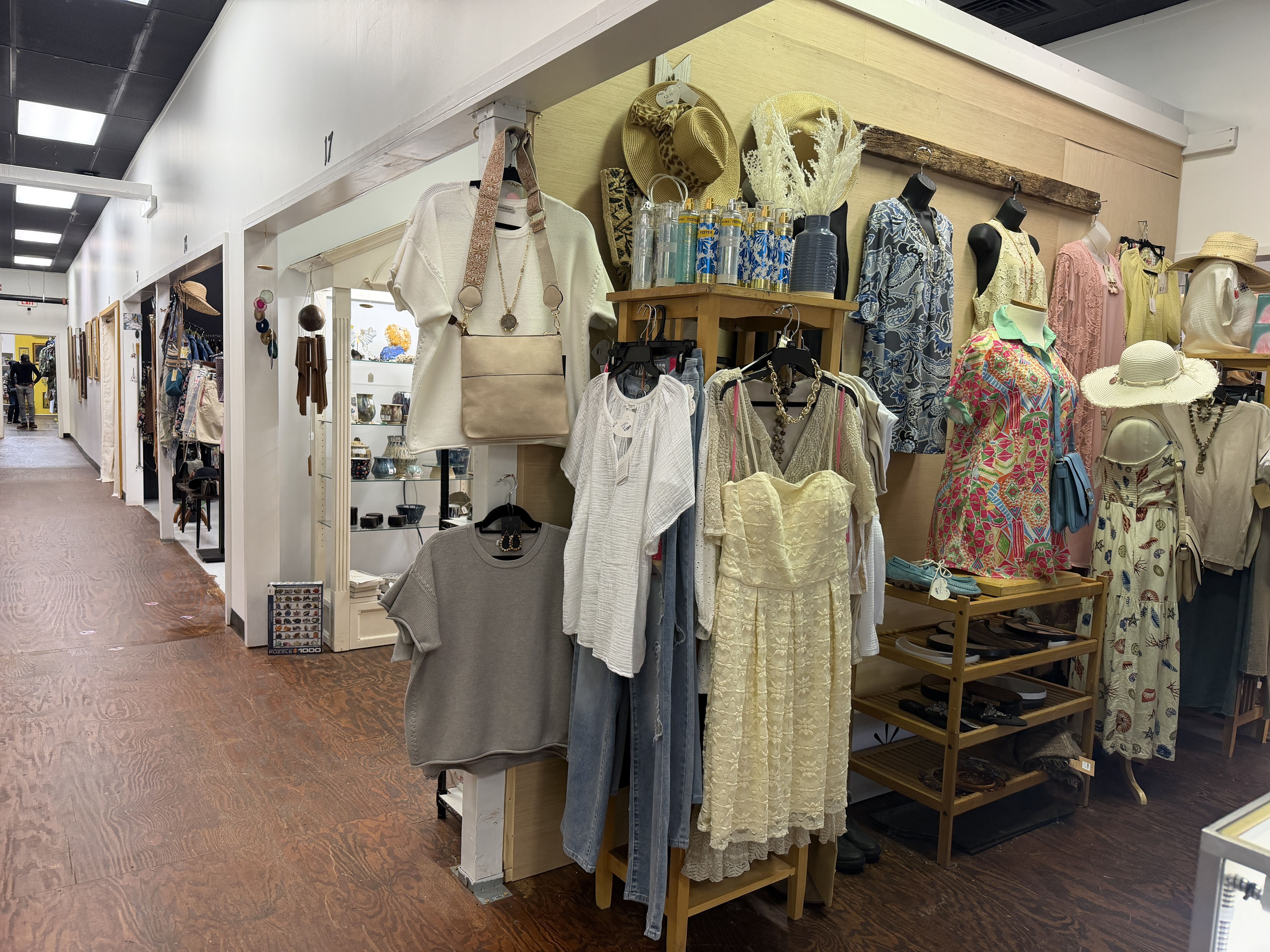 Retail clothing shop with rows of dresses and mannequins in pastel lace outfits; a beige bag on a white sweater in the foreground, and hats, sandals, and accessories on shelves.