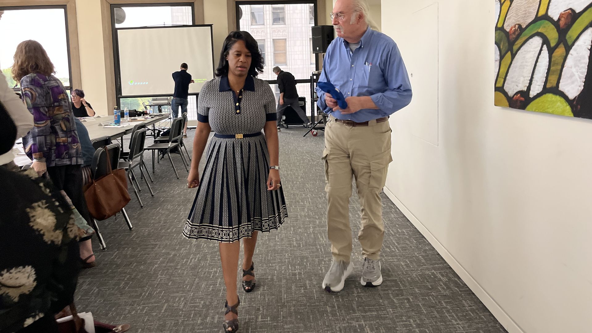 Indoor office or conference room with people walking and sitting around tables; woman in patterned navy dress walks beside man in blue shirt and beige pants holding blue item.
