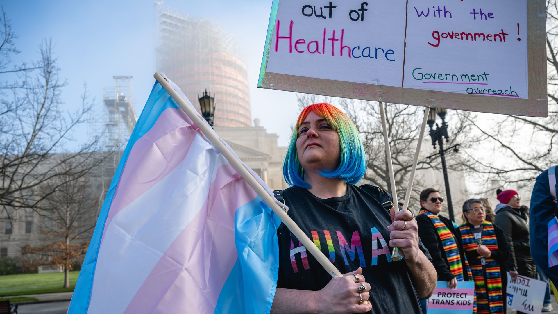 A person with rainbow-colored hair down to the chin hold a blue, pink and white striped flag and a handmade sign.
