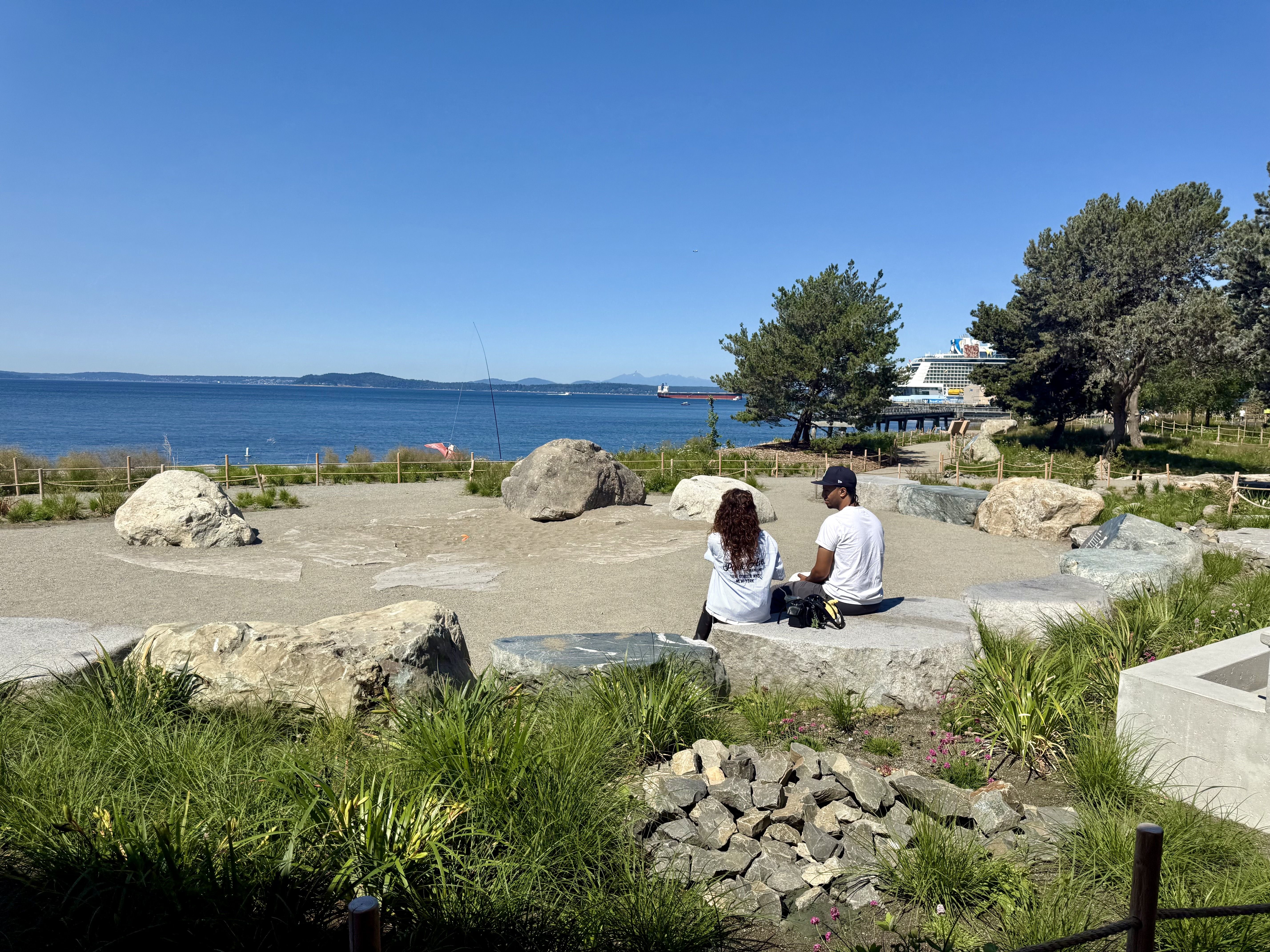 Bright blue sky over calm sea; a distant ship by a pier. In the foreground, large rocks, grasses, and two people in white shirts sitting with their backs to the camera, with trees on the right.