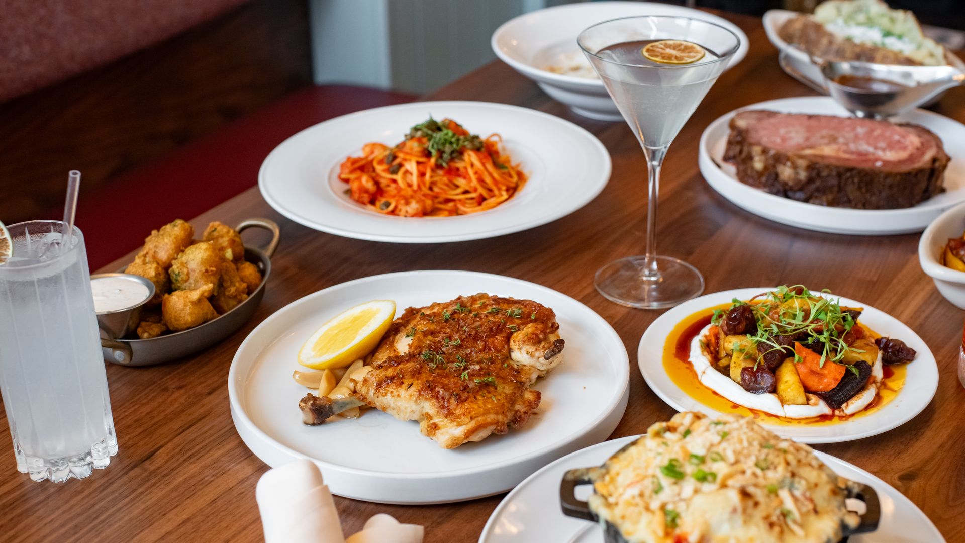 Table with various foods including grilled chicken with lemon, spaghetti in red sauce, mac and cheese, fried appetizers, sliced roast beef, and a clear cocktail with a lemon slice.