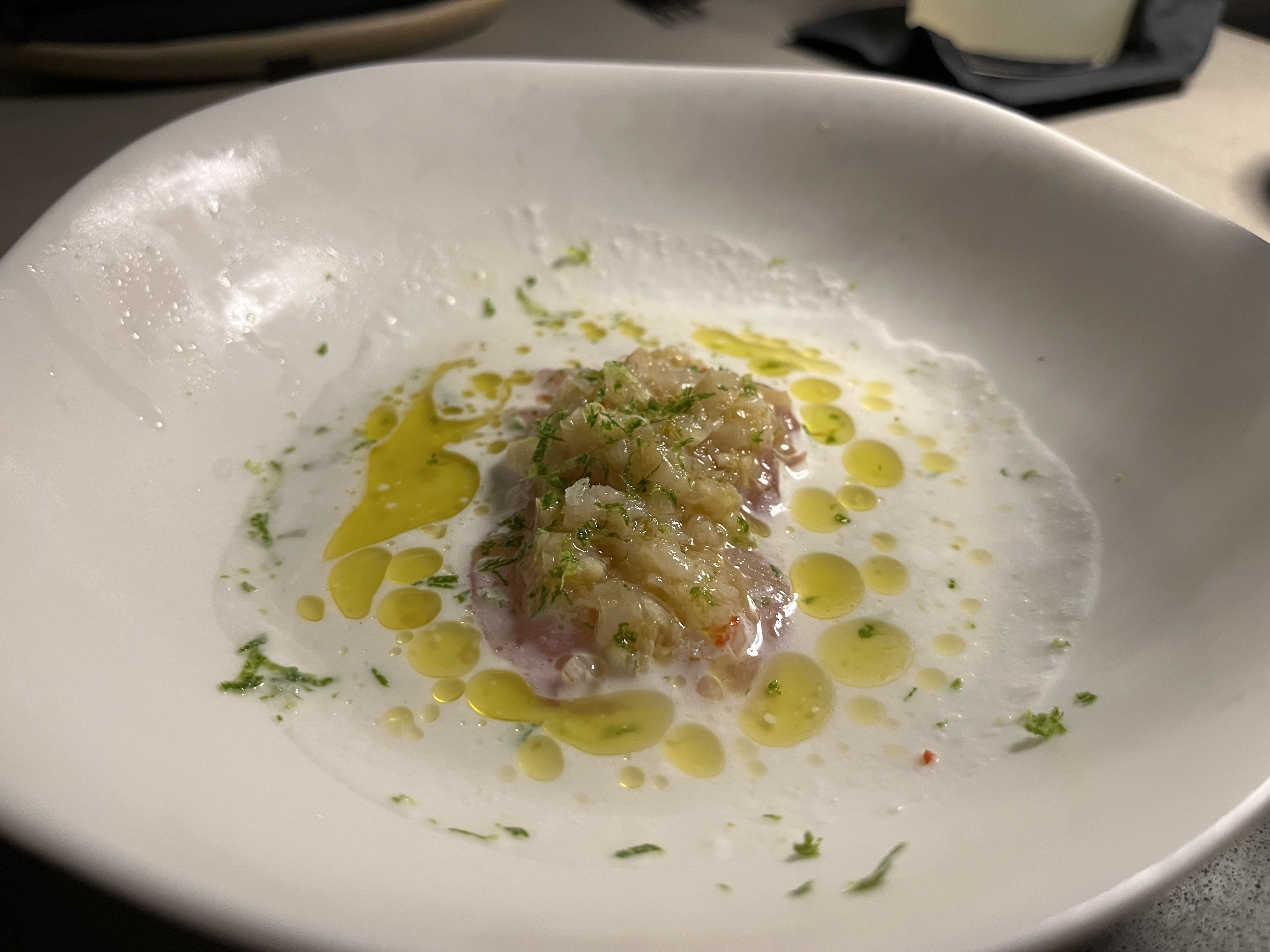 Close-up of a gourmet dish in a white bowl featuring finely chopped seafood topped with green herbs, surrounded by a creamy white sauce with drizzled yellow oil droplets.