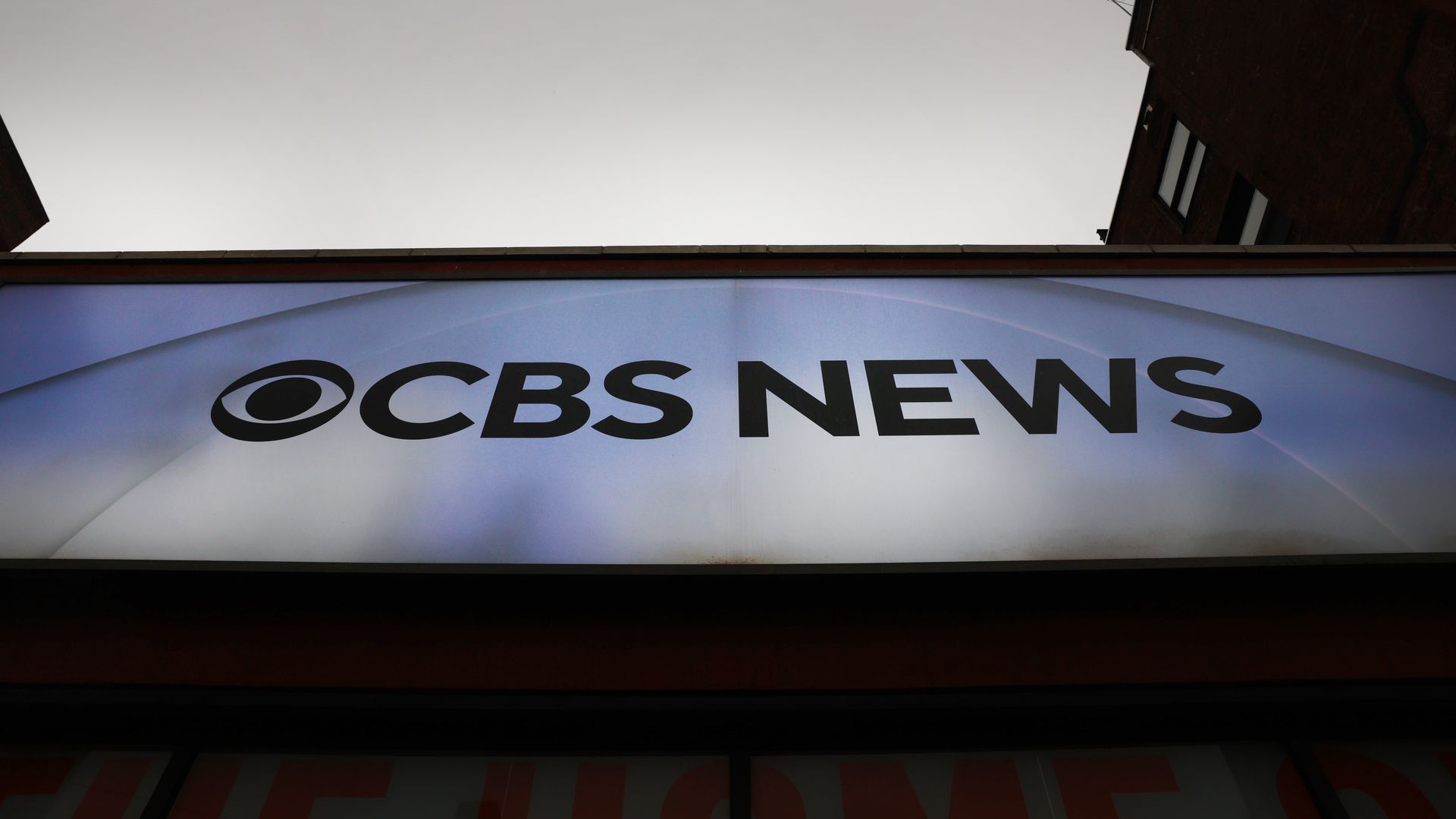 A sign reading "CBS News" with the CBS eye logo on a blue-to-white gradient background, set against a gray sky and surrounding buildings.