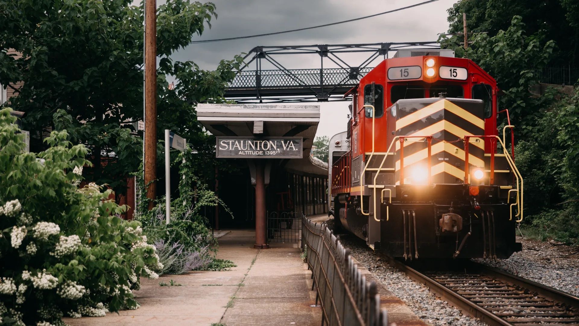 Red and black locomotive with lights on arriving at Staunton, VA train station, surrounded by green trees and bushes under a cloudy sky.