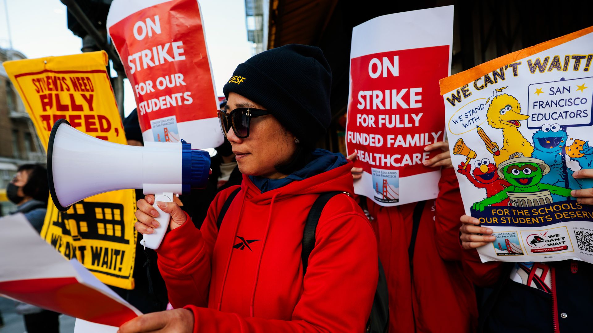 Person in red hoodie and black beanie using megaphone at strike with signs reading "On Strike for Our Students" and "We Can't Wait!" supporting San Francisco educators.