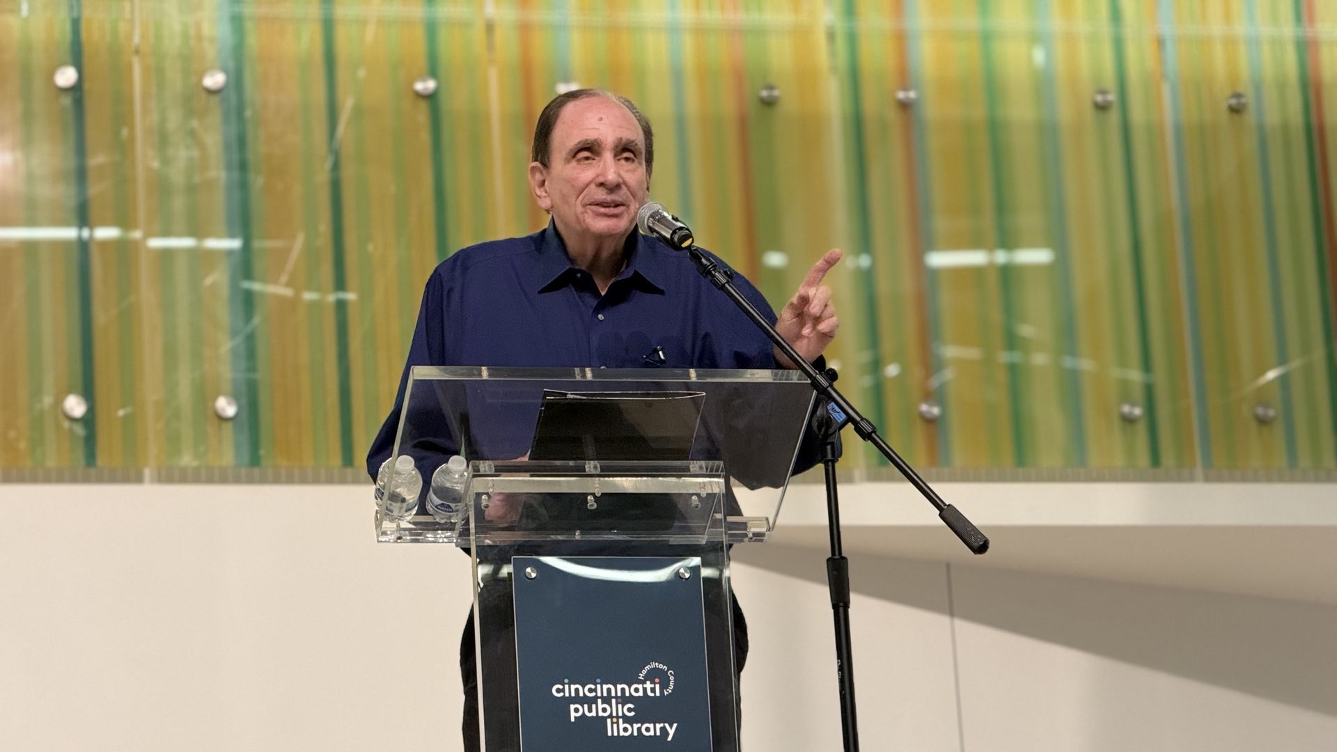 An older man in a dark blue shirt speaks at a clear podium with a microphone; a striped glass wall stands behind him. Water bottles sit on the lectern, Cincinnati Public Library logo visible.