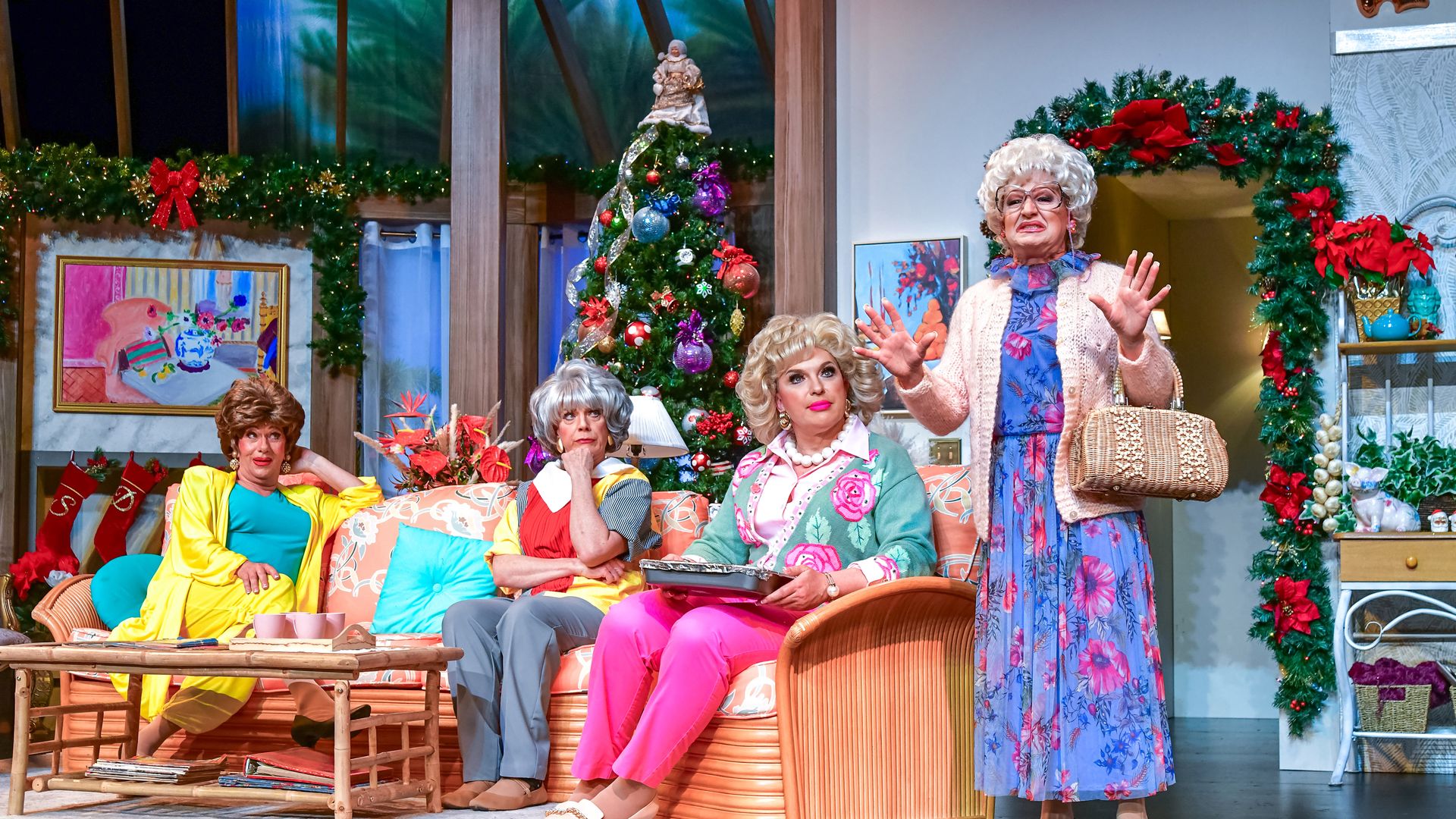 Four women in colorful vintage outfits in a festive living room decorated with a Christmas tree, wreaths, red bows, and stockings, with one woman standing and three seated on a couch.
