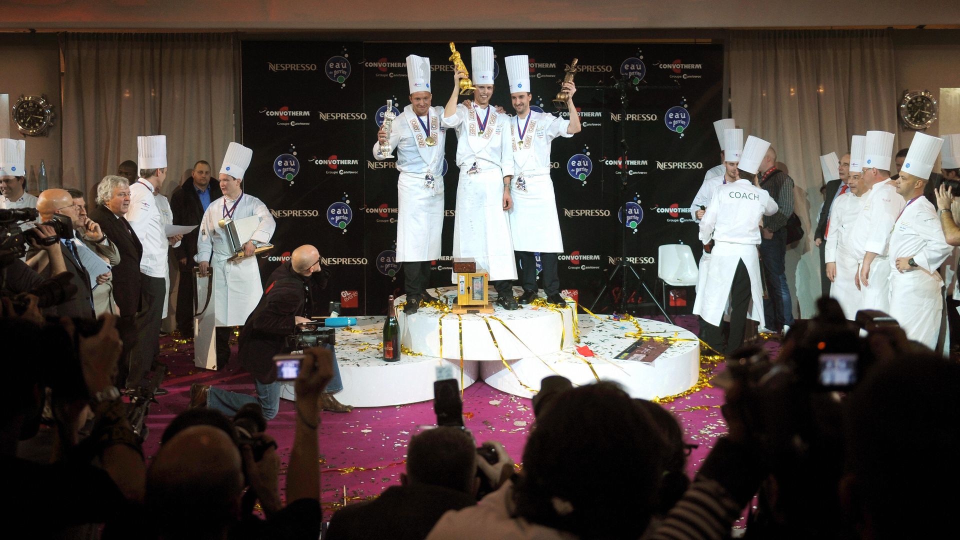A trio of chefs hold trophies while standing on a riser at the center of a crowd of chefs.