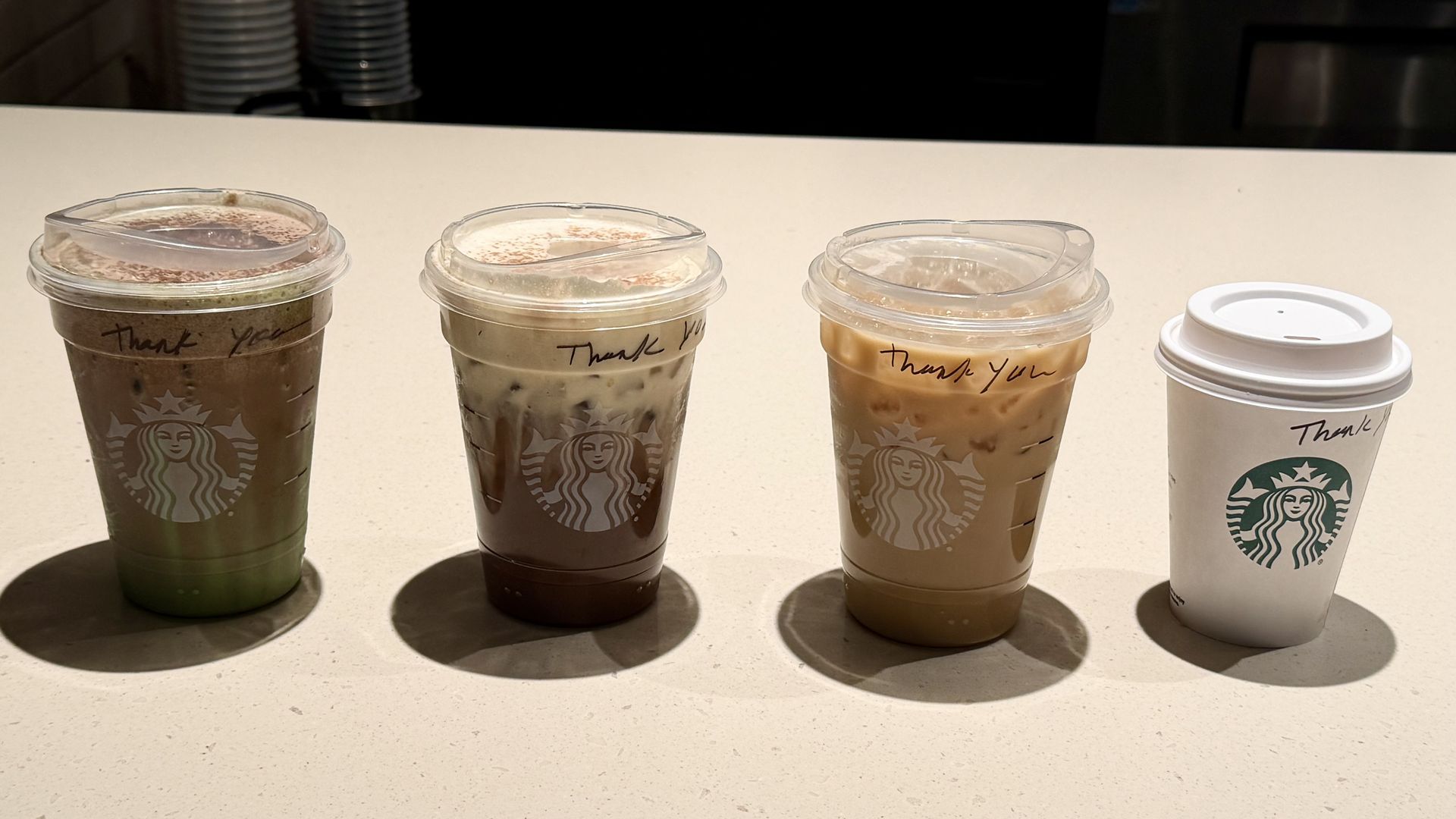 Four Starbucks drinks on a light-colored countertop: three iced beverages in clear plastic cups with "Thank You" written on them, ranging from green to brown to light tan, and one hot drink in a white cup with a lid.