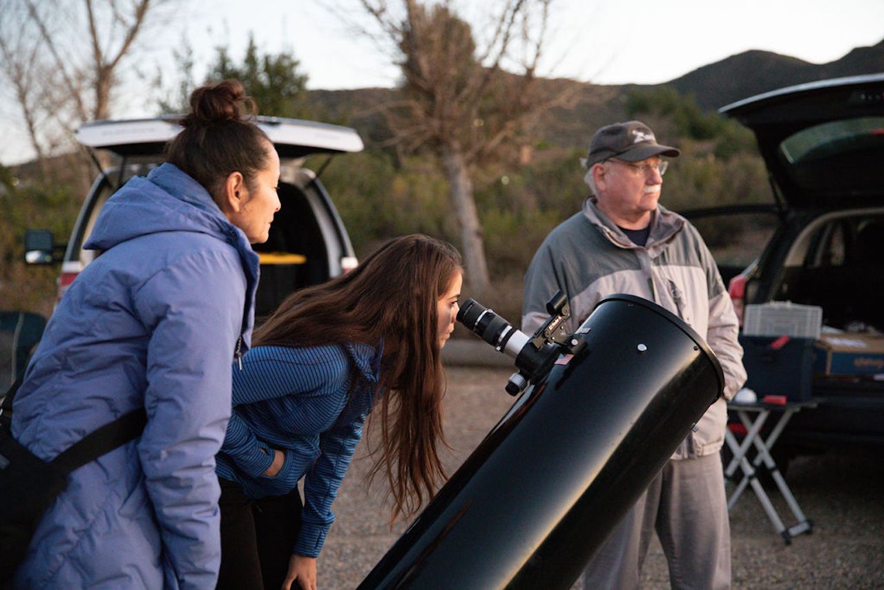 Three people outdoors around a large black telescope: a woman peers into the eyepiece, another person watches, and an older man stands by an open car trunk with hills in the background.