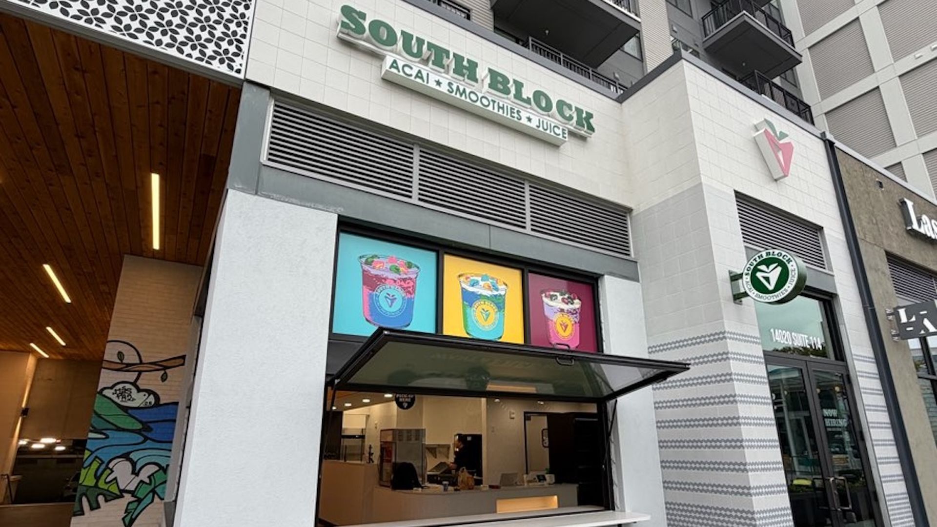 Exterior of South Block smoothie and juice bar with white walls, three stool seats, and colorful smoothie illustrations above the order window on a gray patterned sidewalk.