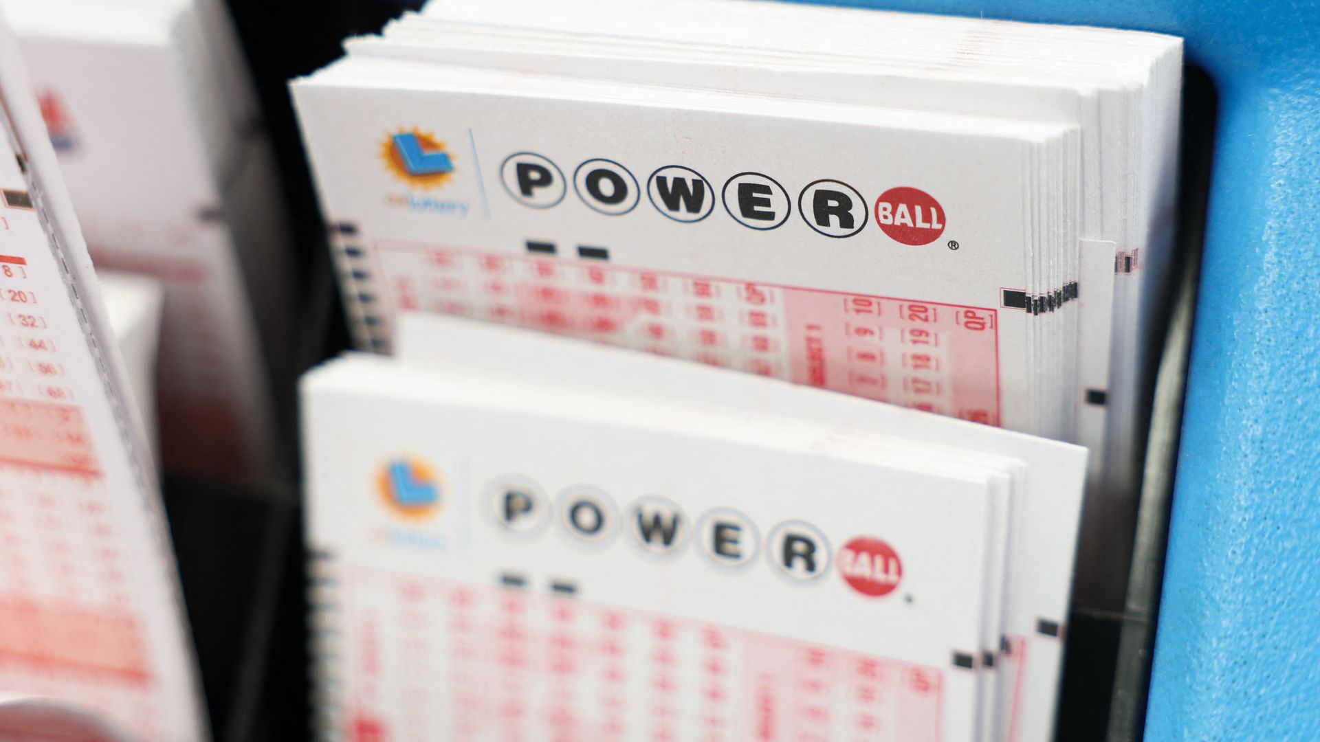 Close-up of blank white Powerball lottery tickets with black and red lettering, held in a blue ticket dispenser.
