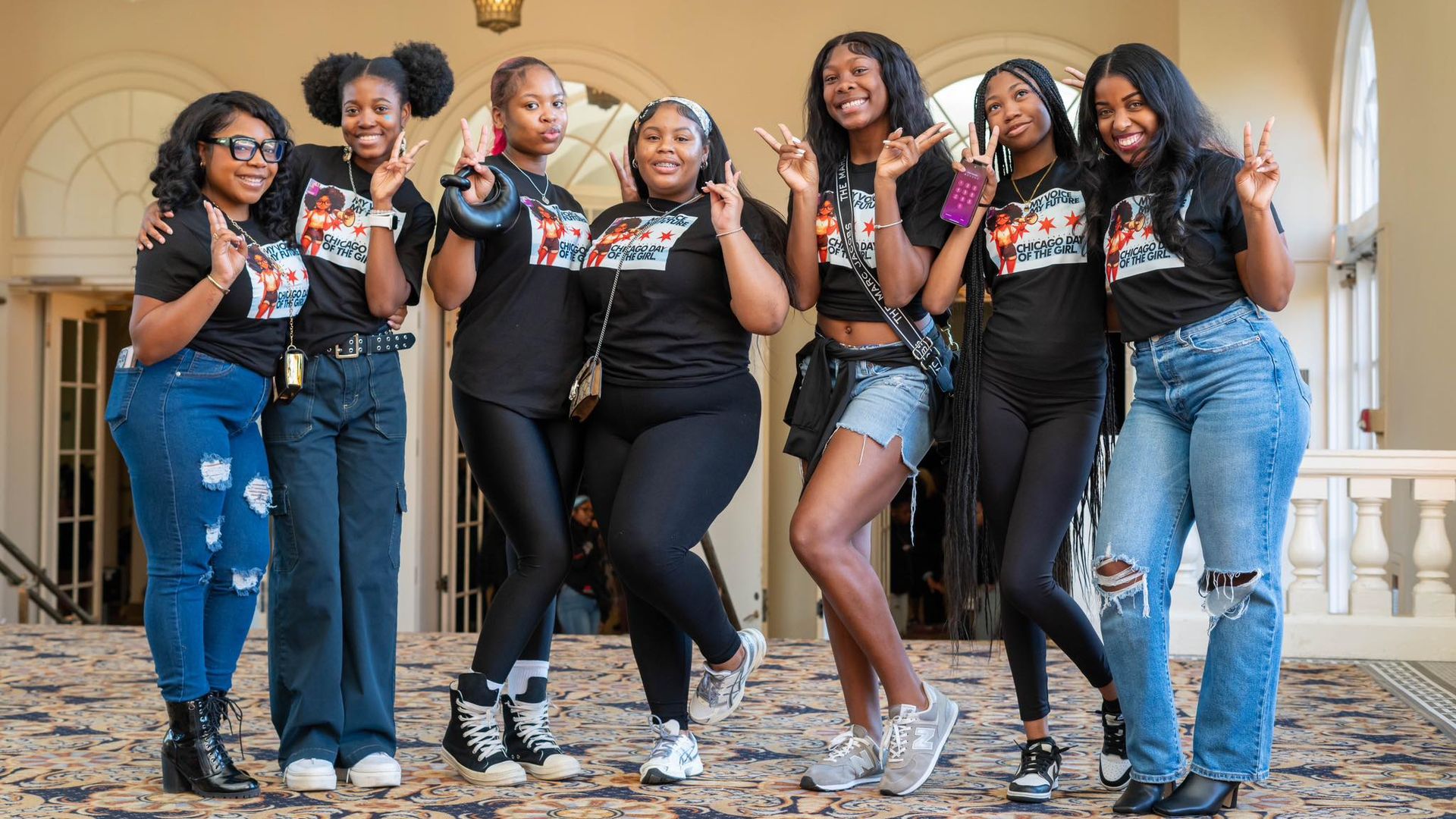 Seven smiling young women in a bright hallway wear matching black t-shirts with a colorful print that reads "Chicago Day of the Girl" and show peace signs with their hands.