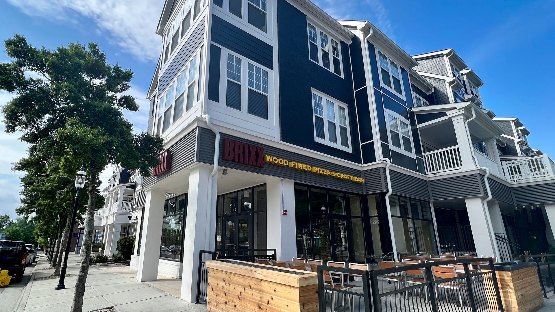 Corner view of a blue and white building with a BRIXX Wood Fired Pizza & Craft Bar sign, outdoor seating with wooden barriers, a tree-lined street with parked cars on a sunny day.