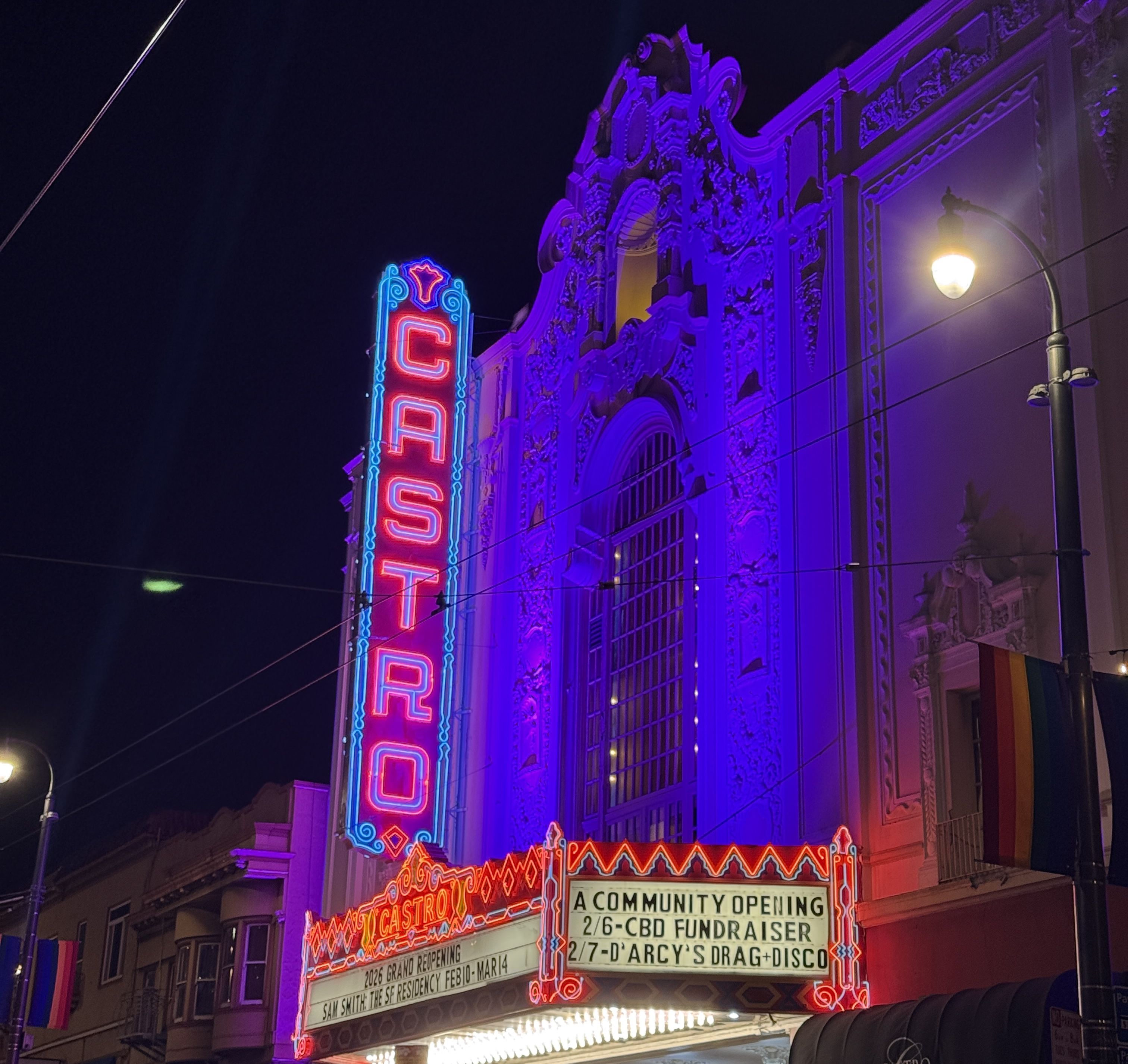An nighttime shot of the exterior of the Castro Theatre with the "Castro" sign lit in red and blue neon, and a lit marquee showing upcoming shows.
