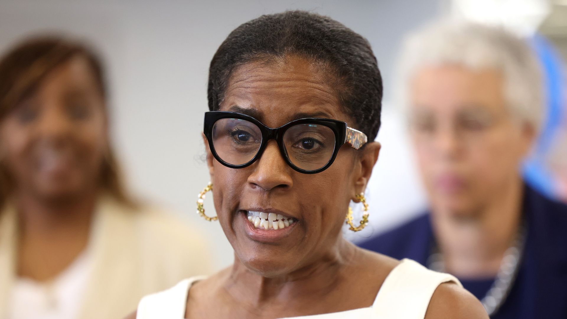 A smiling woman wearing black round glasses, gold hoop earrings, and a white sleeveless dress, speaking in front of a blurred group of people.