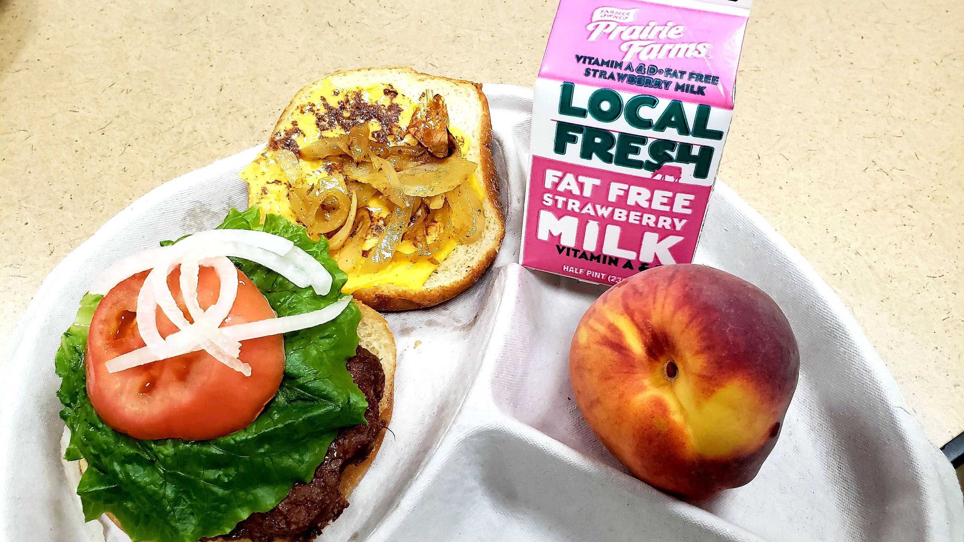 Open-faced burger with lettuce, tomato, and onions on paper plate next to peach and small carton of milk.