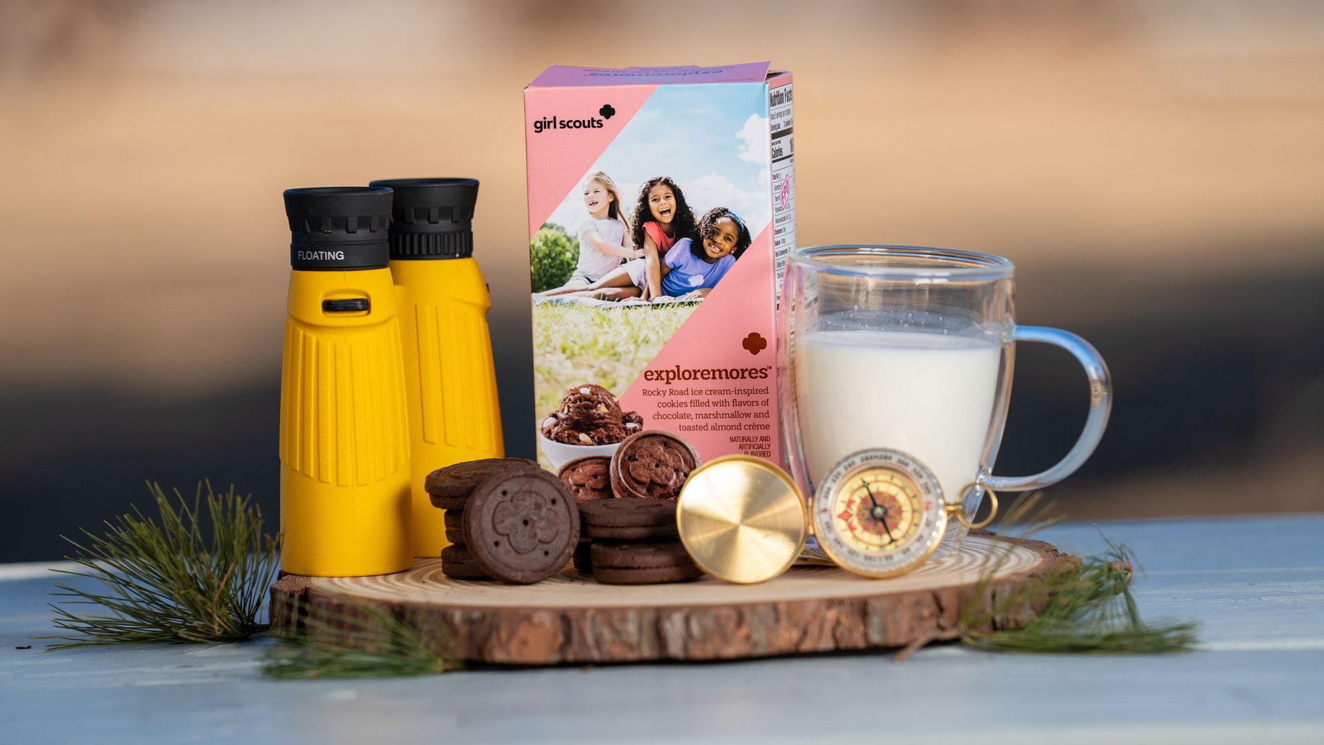Yellow binoculars, Girl Scouts "exploremores" cookie box, chocolate cookies, brass compass, and glass mug of milk on a wooden slab outdoors.