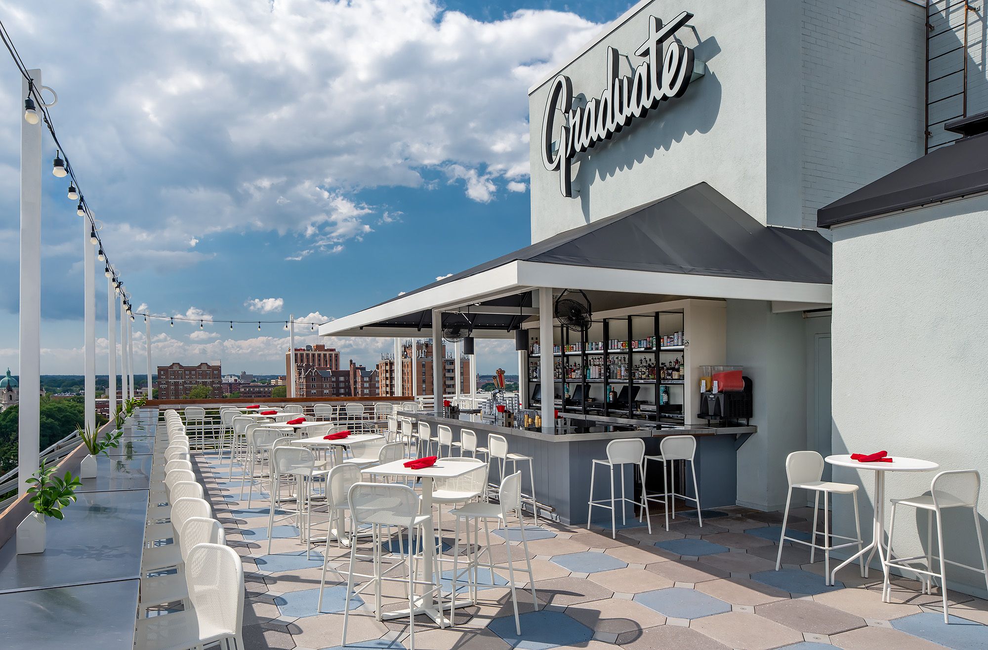 Rooftop bar with white tables and stools, red napkins, and a long counter under a canopy. A large "Graduate" sign on a gray wall, string lights, and a distant city skyline under a blue sky.