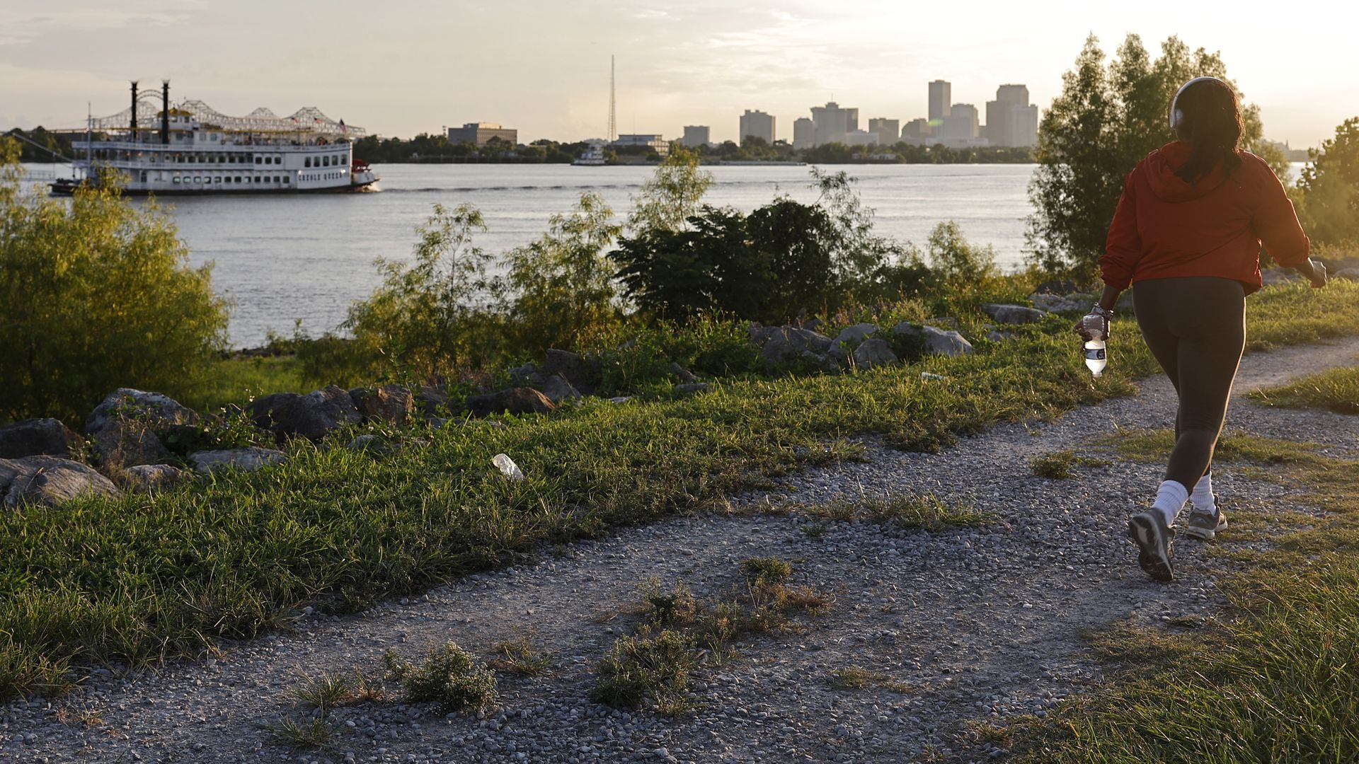 Person in red jacket walking on gravel path near water at sunset, holding a water bottle, with a city skyline and a riverboat in the background.