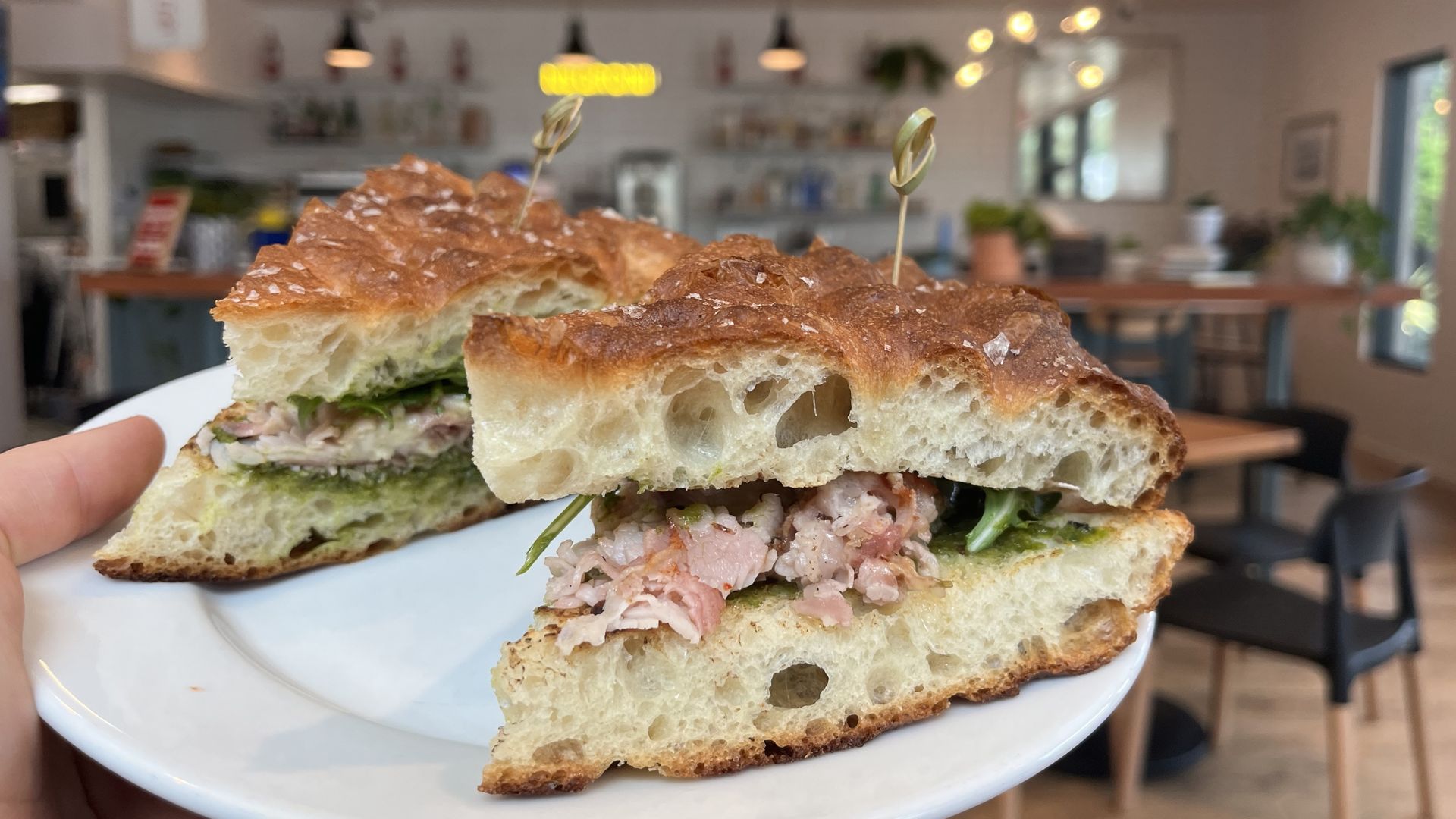 Hand holding a white plate with two focaccia sandwiches filled with sliced meat, greens, and sauce, inside a cozy café with wooden furniture and soft lighting.