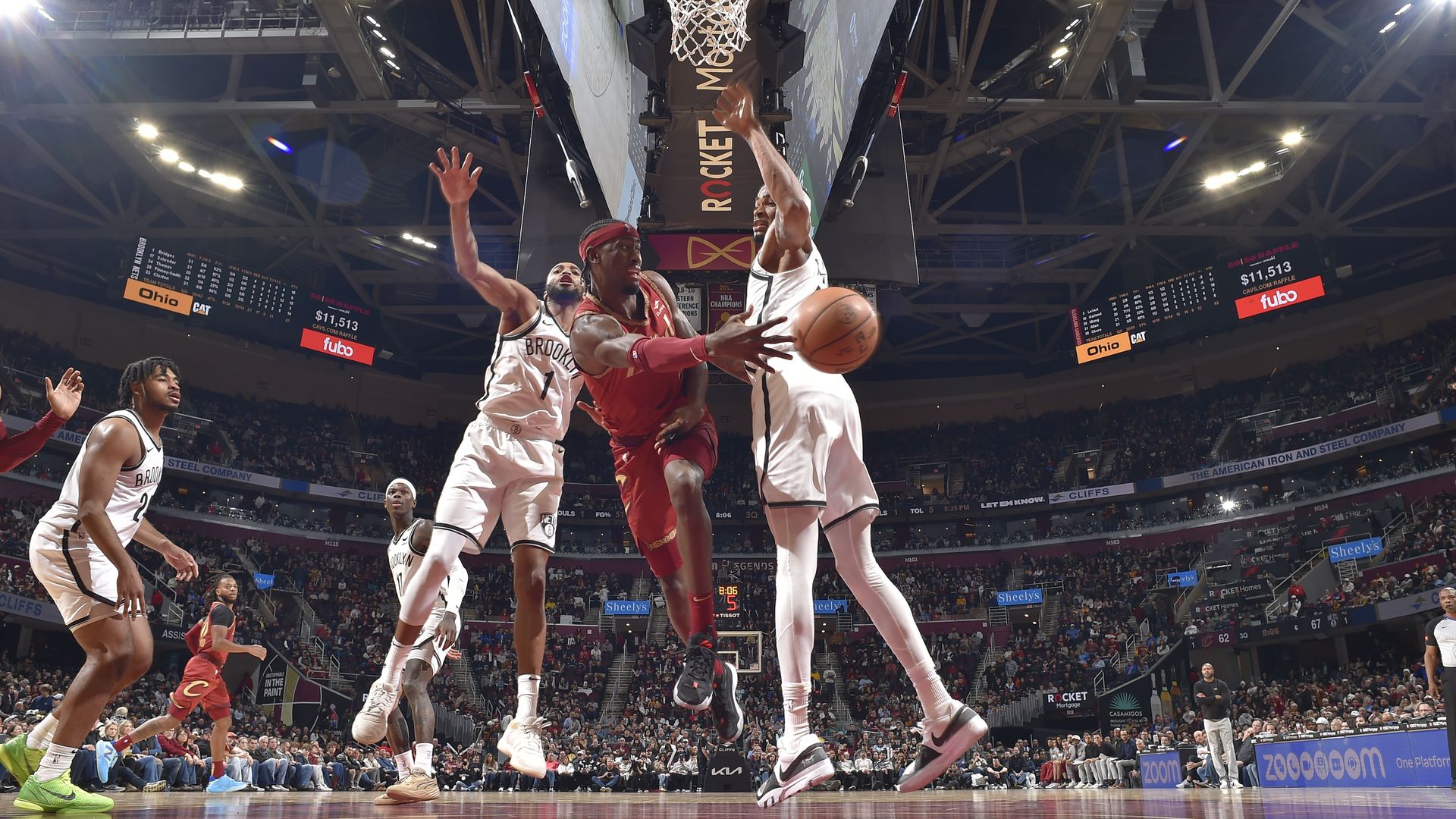 Caris LeVert (red jersey) delivers pass under basketball hoop between two Brooklyn Nets (white jerseys)