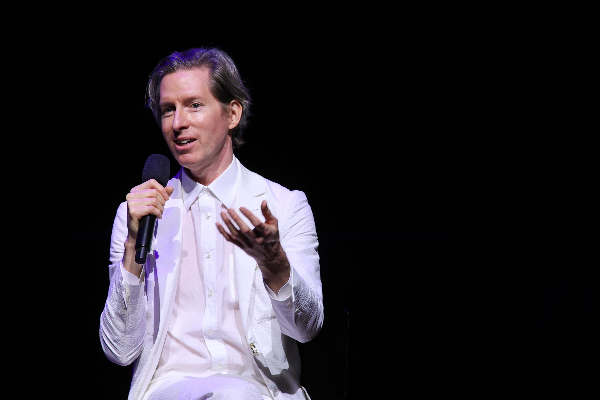 Photo of a man in a white shirt and suit in a black background. He's holding a mic. 