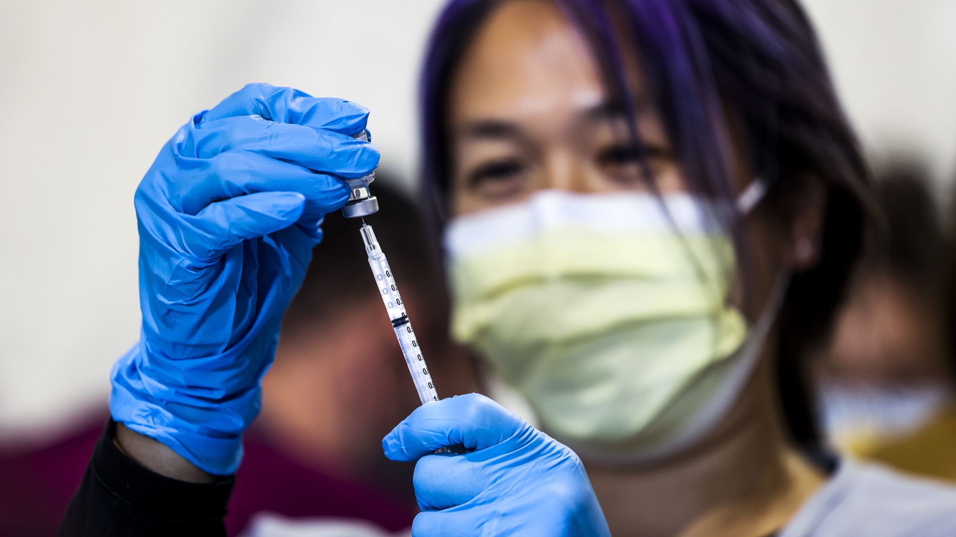 A woman holds a syringe while wearing gloves and a face mask 