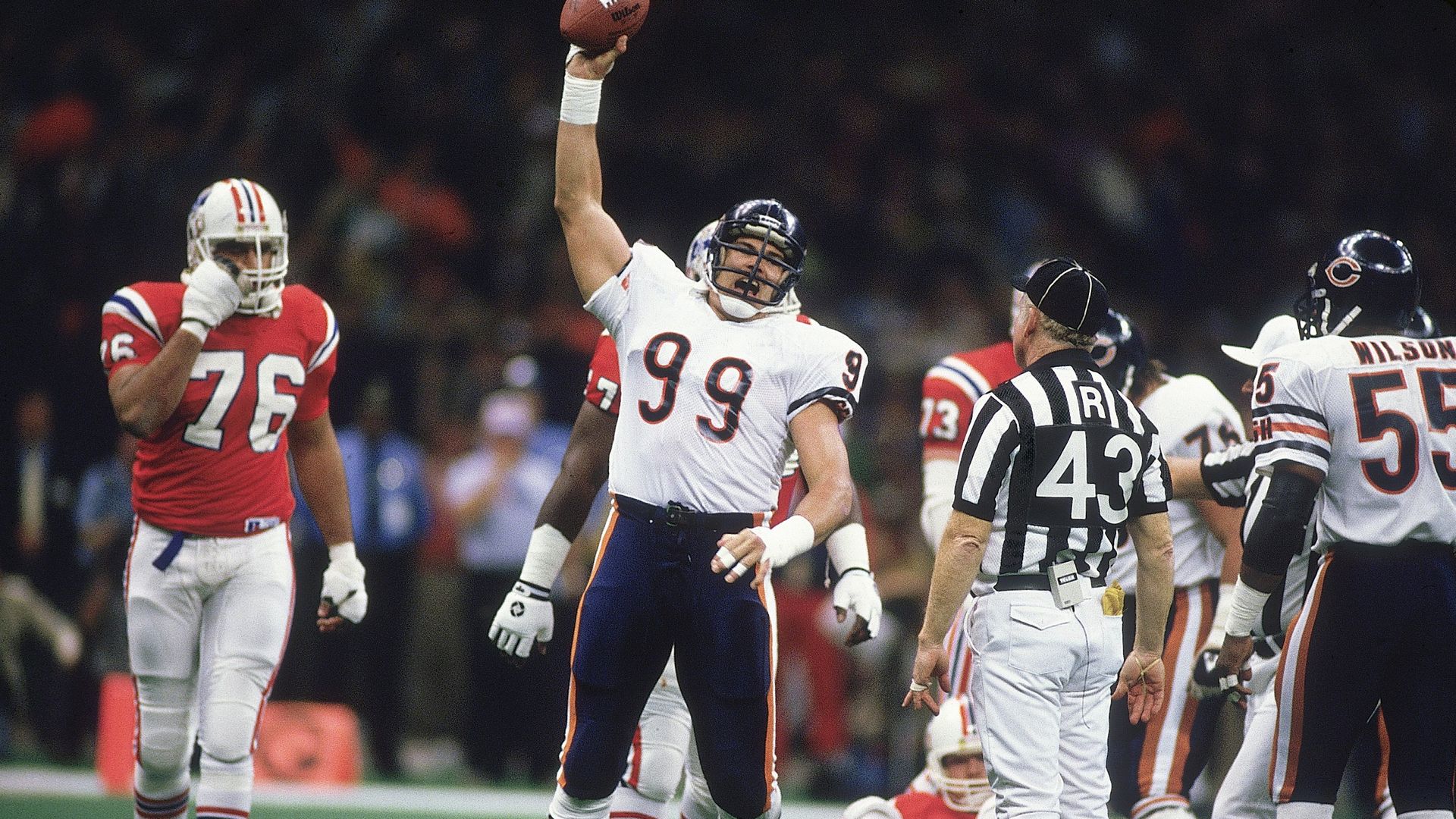 Football player in white jersey 99 raises ball during game, surrounded by teammates and opponents in red, with referee in black and white stripes nearby on green field.