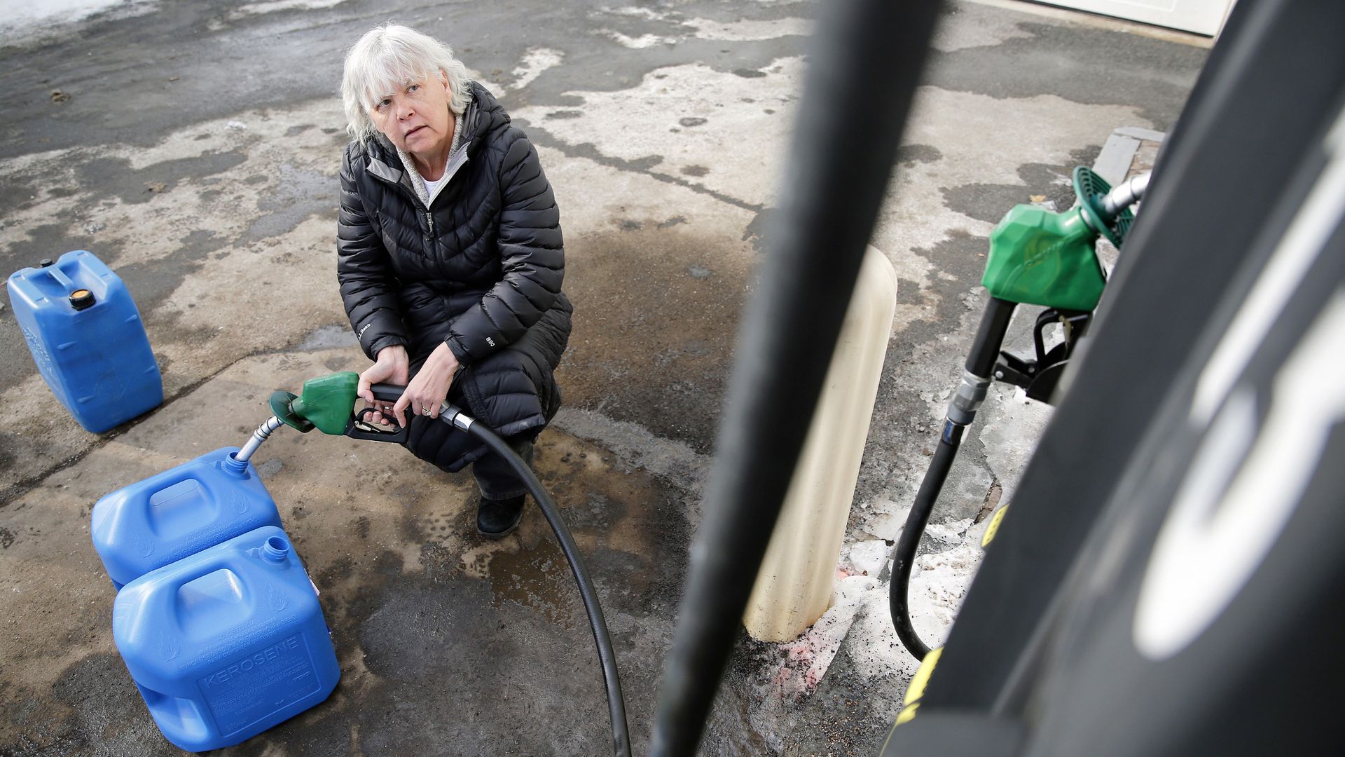 Lady with white hair crouches down filling three blue gasoline jugs with gasoline. 