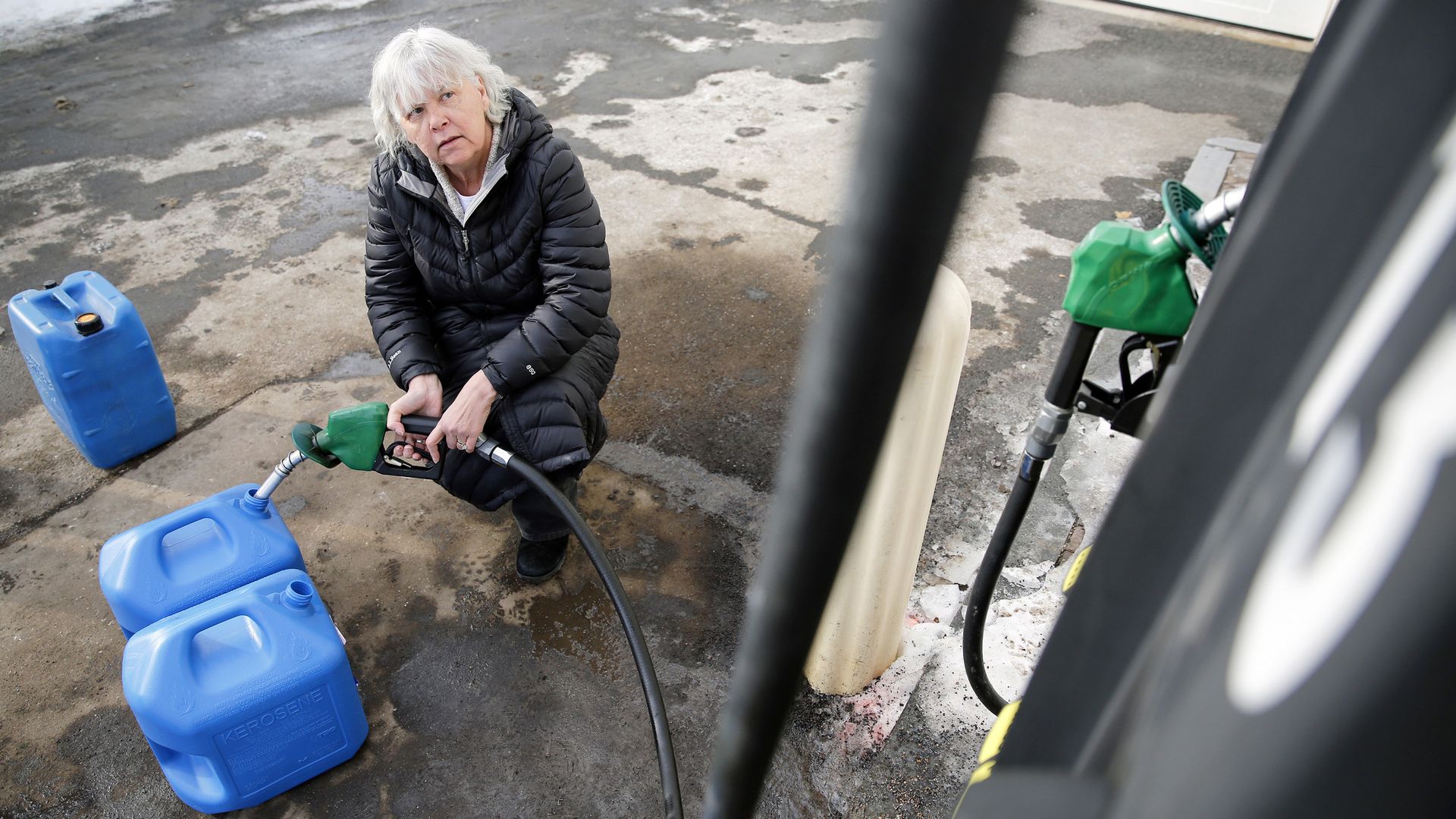 Lady with white hair crouches down filling three blue gasoline jugs with gasoline.