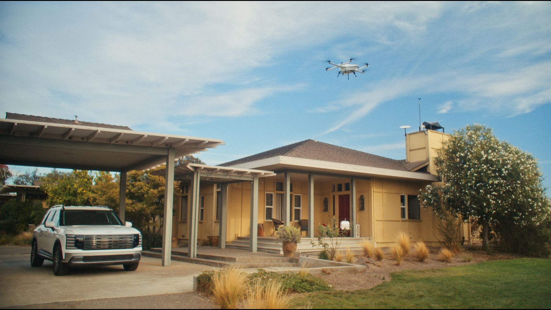 White pickup under a carport beside a beige, single-story house with a brown roof. A white drone hovers in a blue sky above, with wispy clouds; dry landscaping and a flowering tree on the right. The drone is lowering a white package on a tether.