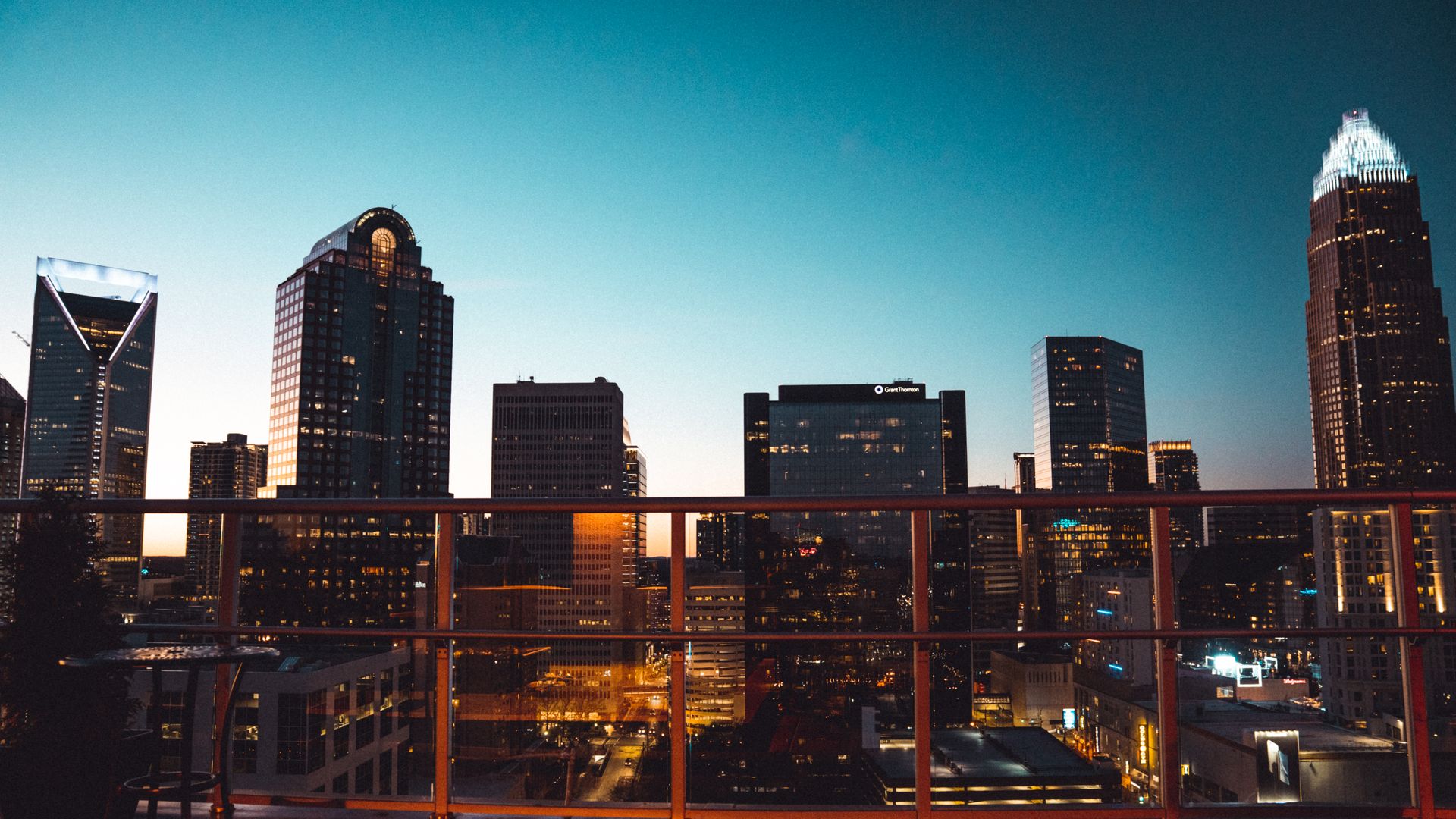 City skyline at dusk with a blue gradient sky, a left glass building with a V-shaped top, a central rounded-topped tower, and a right building with a bright crown; orange railing in the foreground.