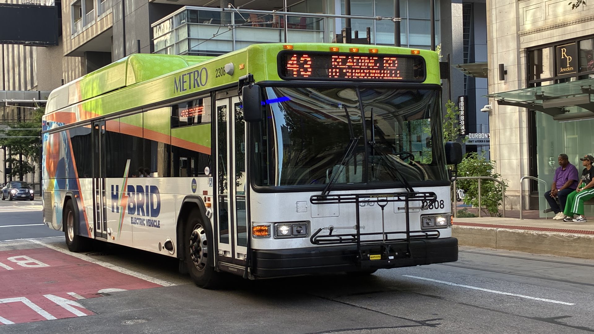 Bright lime-green and white city bus with a rainbow stripe, METRO HYBRID, on a busy street. Route 43 digital sign glows; two people sit at a bus stop to the right.