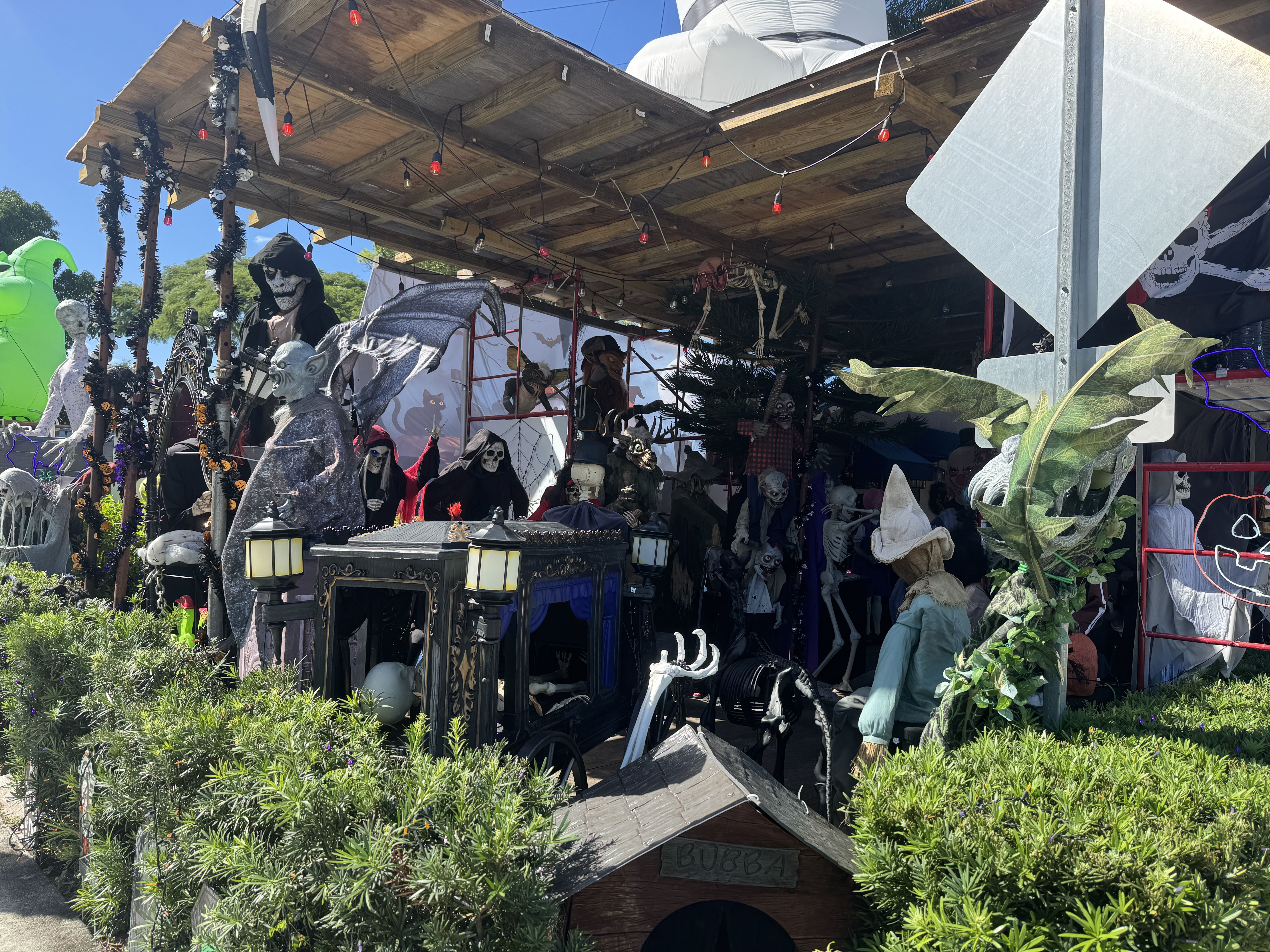 A Halloween display on a wooden open shack; numerous skeletons and grim reapers, skulls and props hang from the ceiling and around the stall, with string lights, cobwebs, and greenery in front.