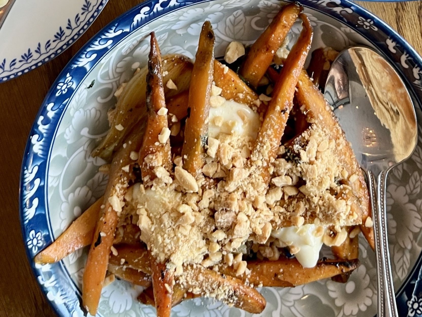 Blue and white floral patterned bowl filled with roasted orange baby carrots topped with crumbled nuts and creamy dollops, beside a silver spoon on a wooden surface.