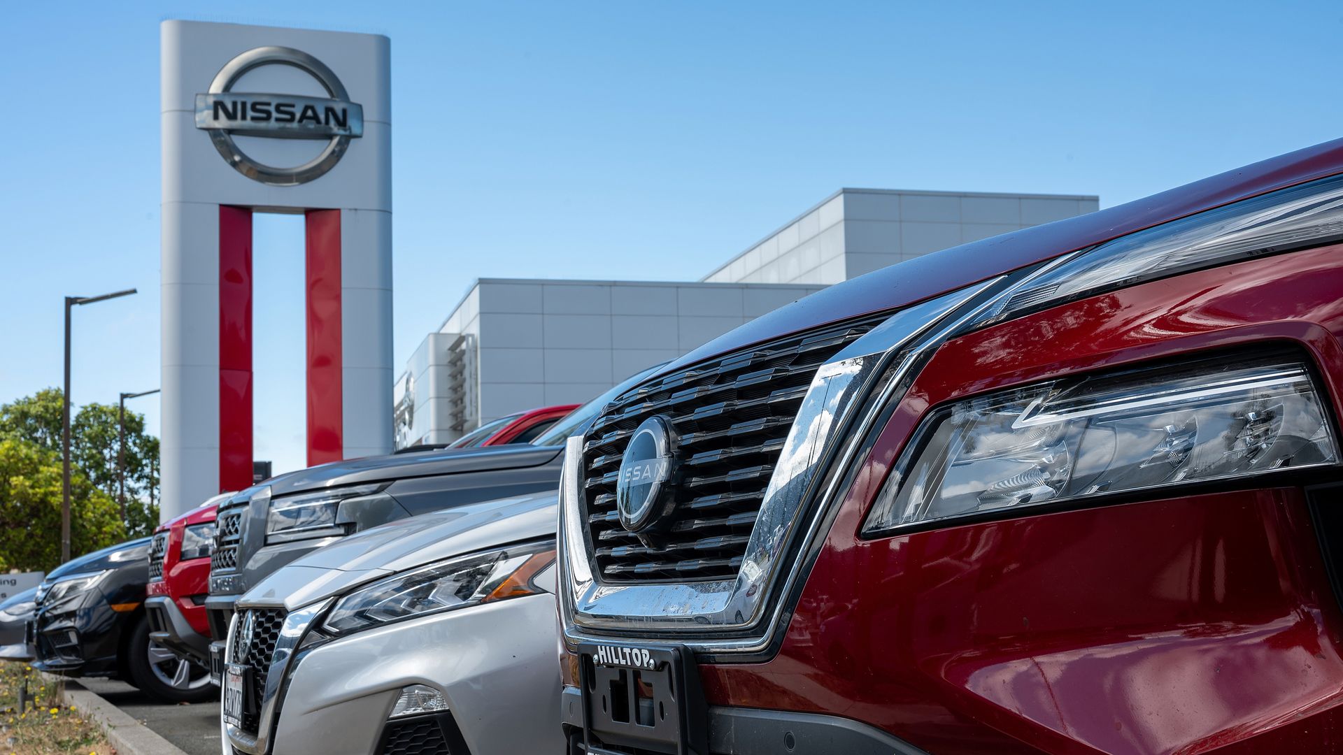 Row of new Nissan vehicles at a sunny dealership, foregrounded by a red SUV with chrome grille; a tall Nissan sign with red stripes and a modern showroom behind.